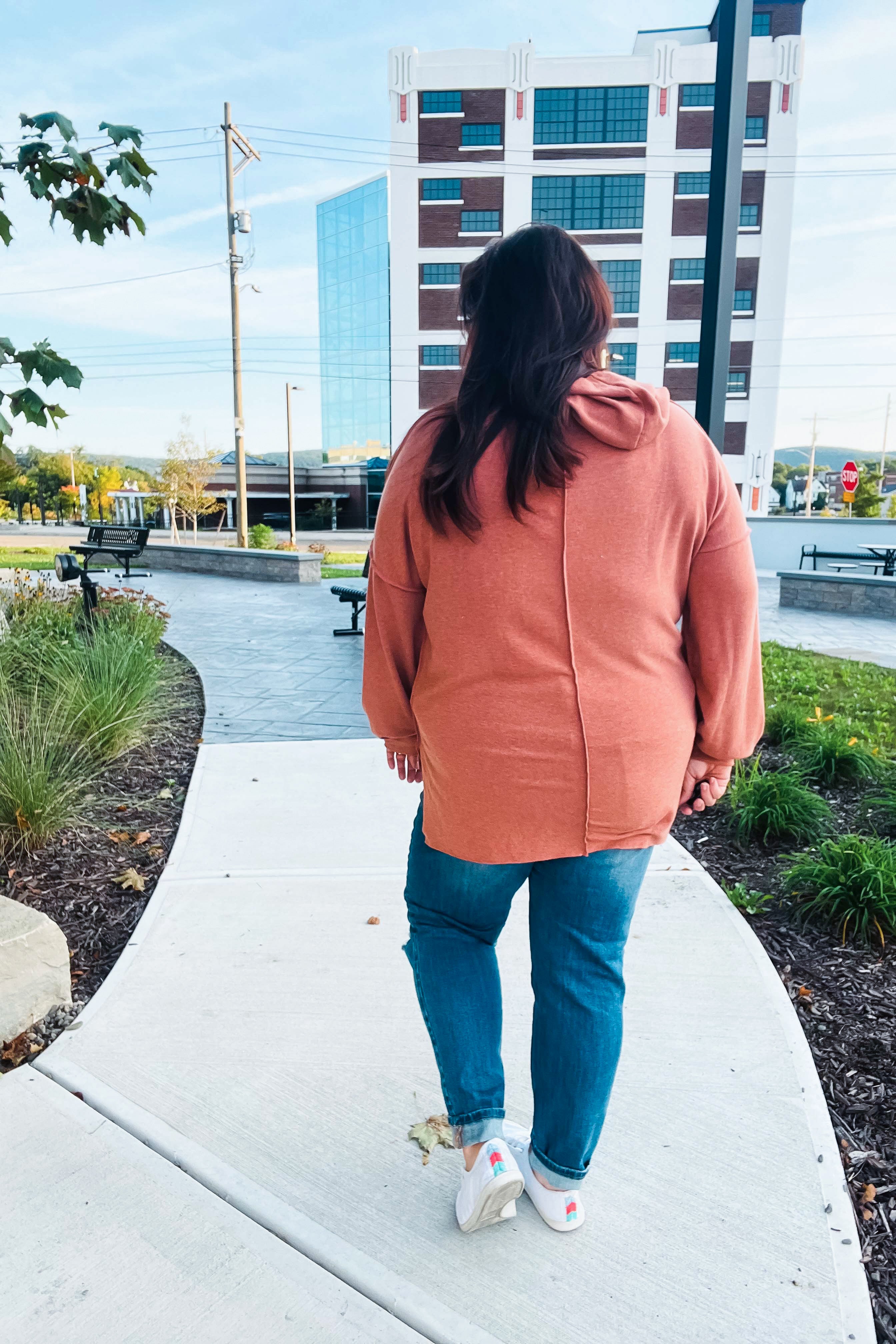 Woman wearing a loose rust-colored hoodie walking on a curved sidewalk in an urban outdoor area