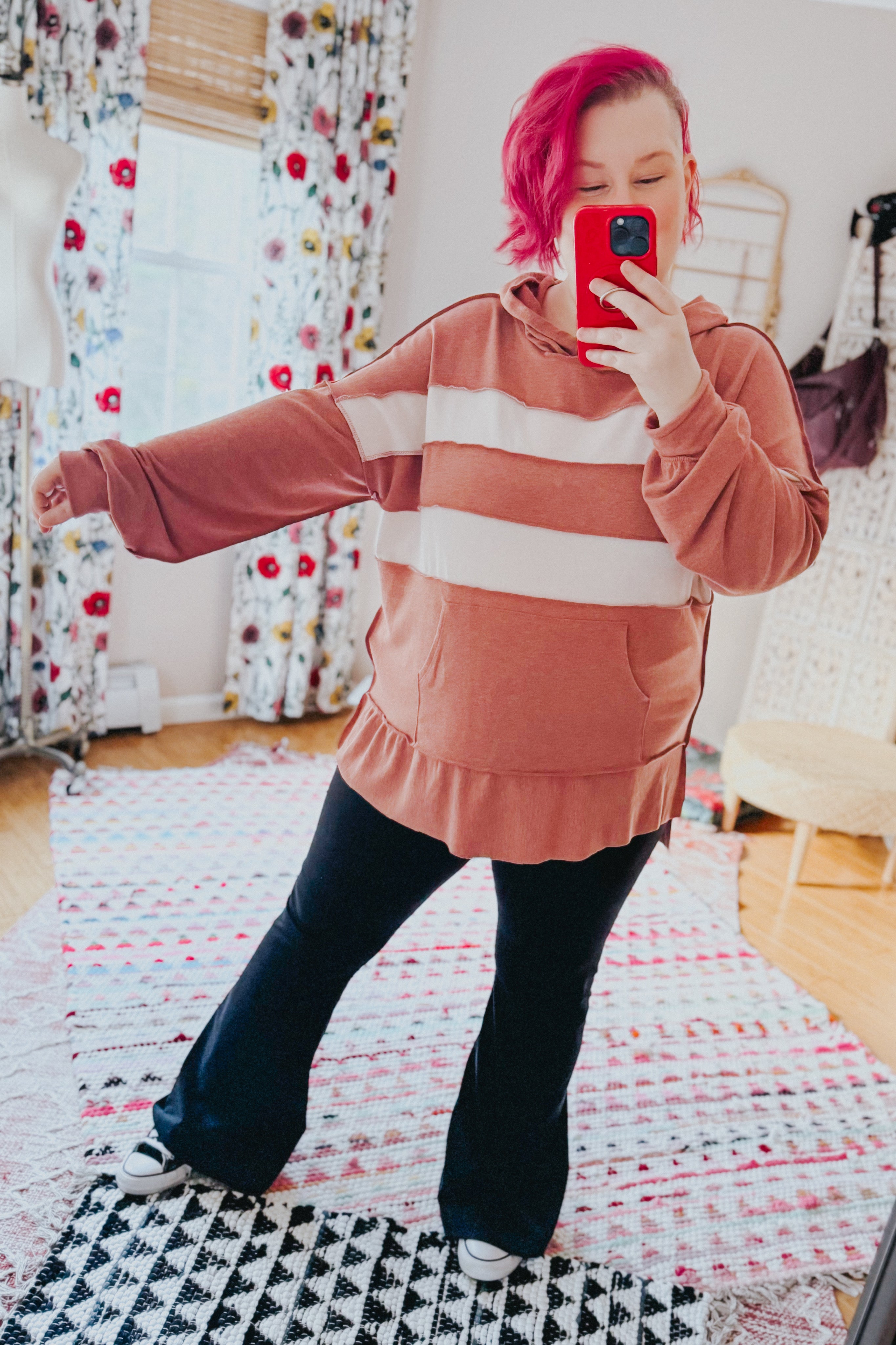 Woman with bright pink hair wearing a loose rust hoodie with wide cream stripes and black pants taking a mirror selfie indoors