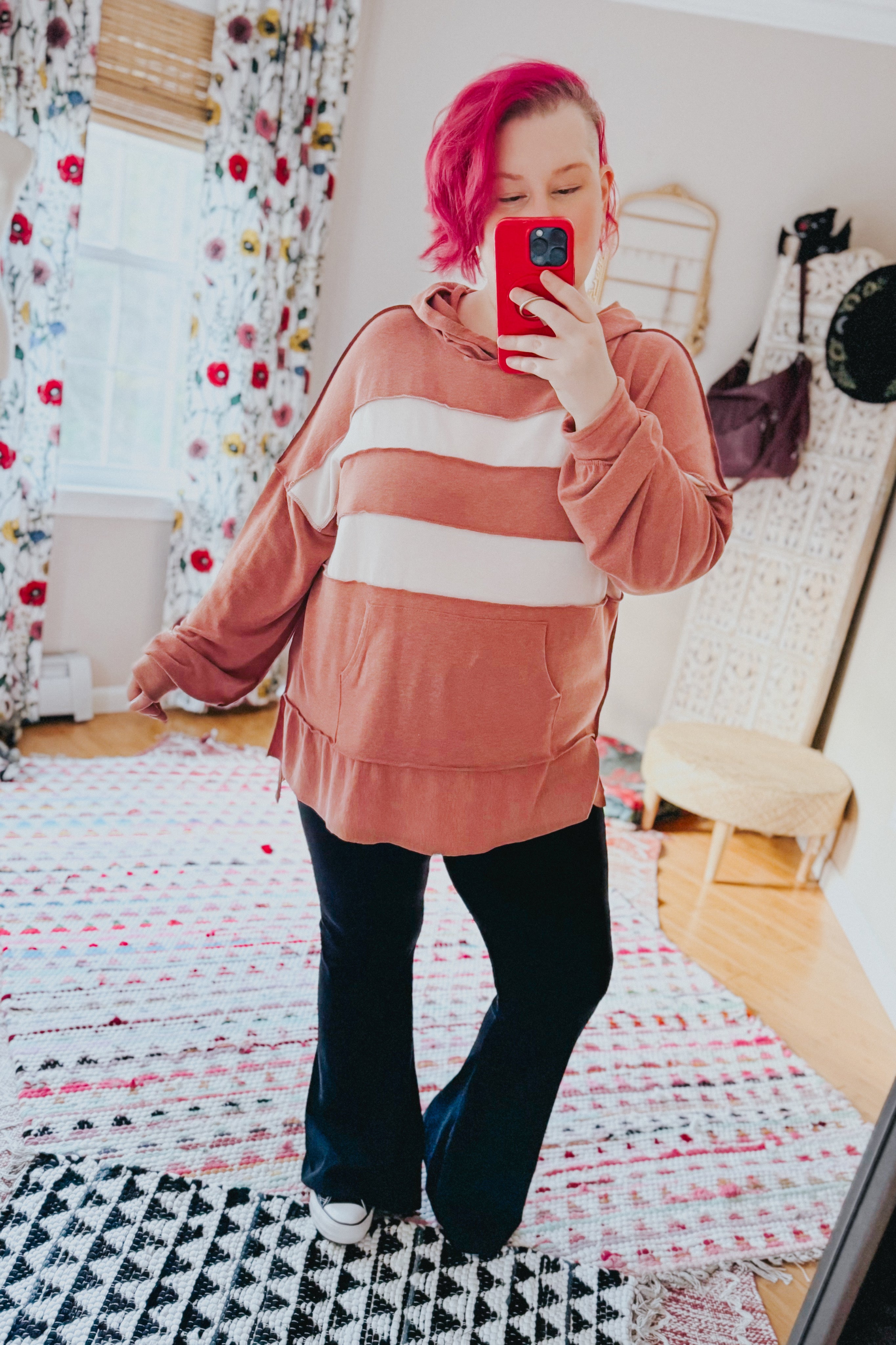 Woman with bright pink hair wearing a loose rust-colored hoodie with wide cream stripes and black pants taking a mirror selfie indoors