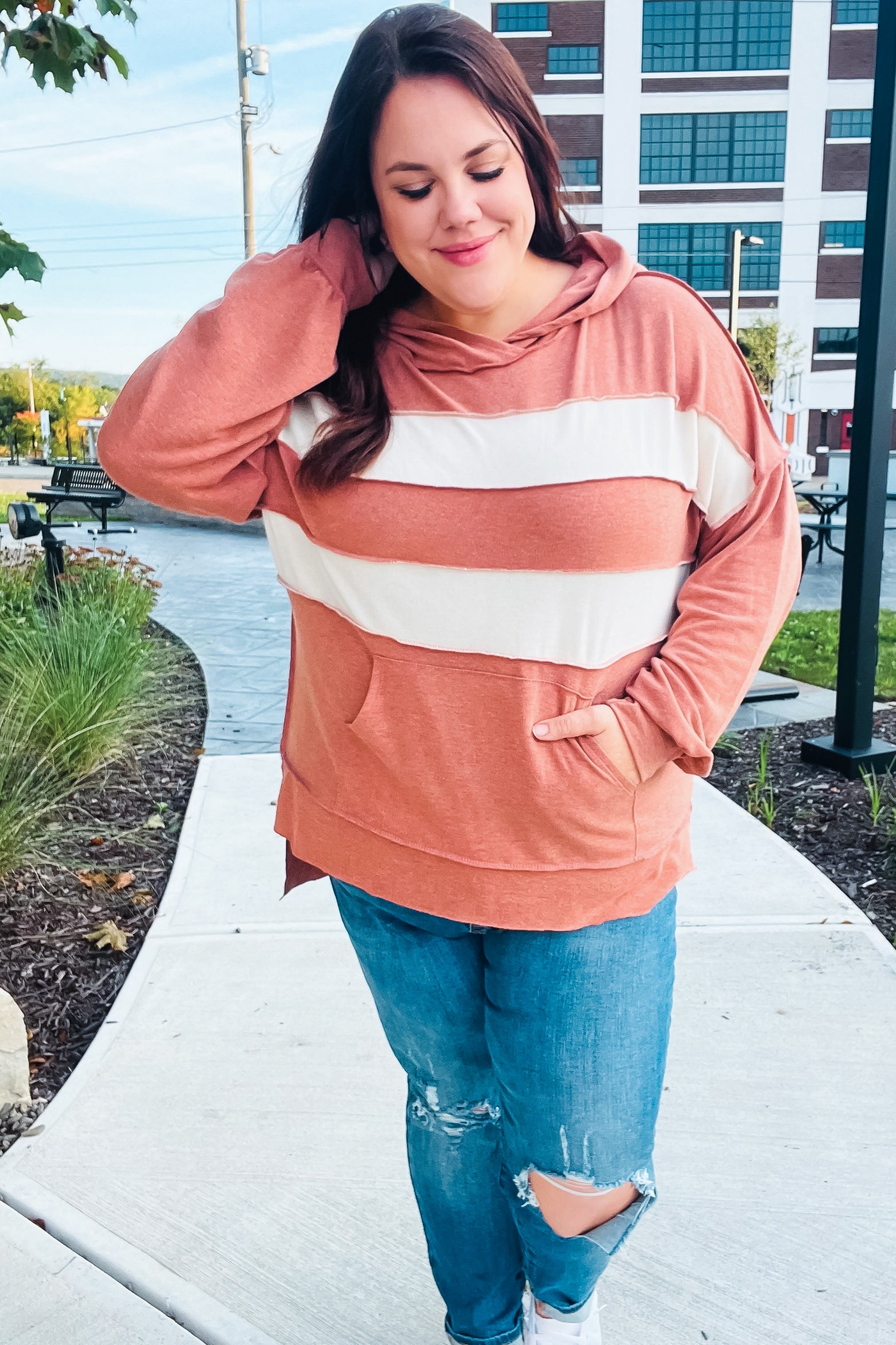 Woman wearing a rust-colored hoodie with wide cream horizontal stripes and ripped blue jeans standing outdoors on a sidewalk