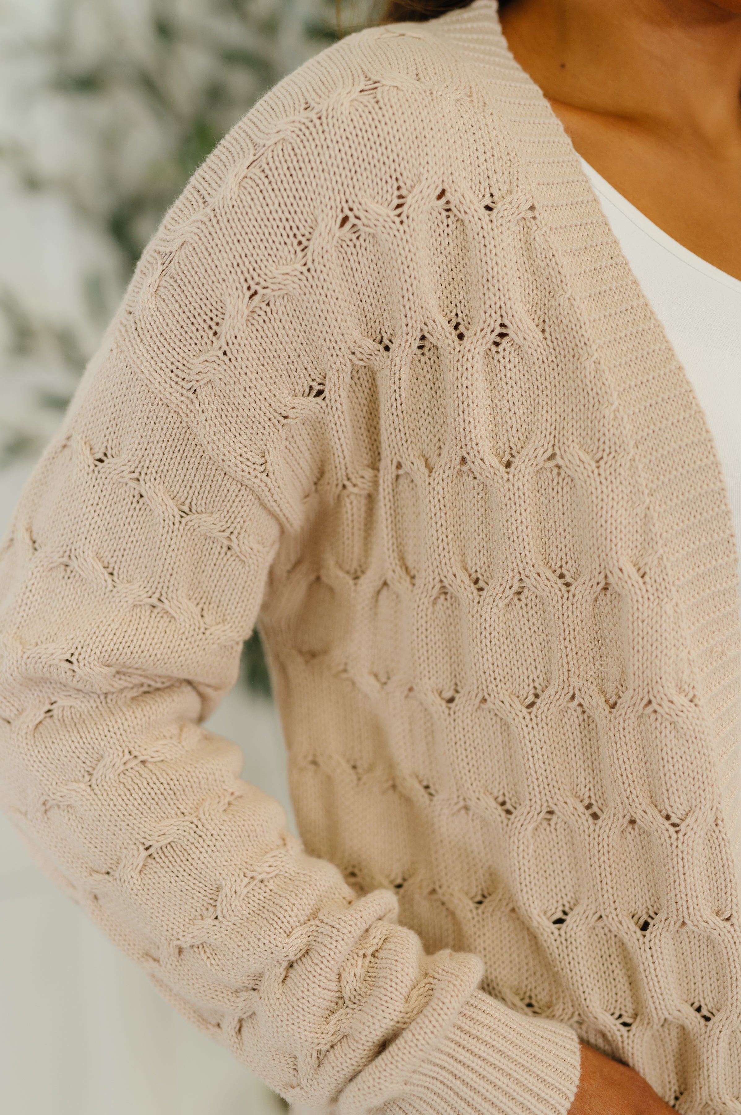 Close-up of beige airy cable knit cardigan sleeve and textured pattern worn over white top indoors