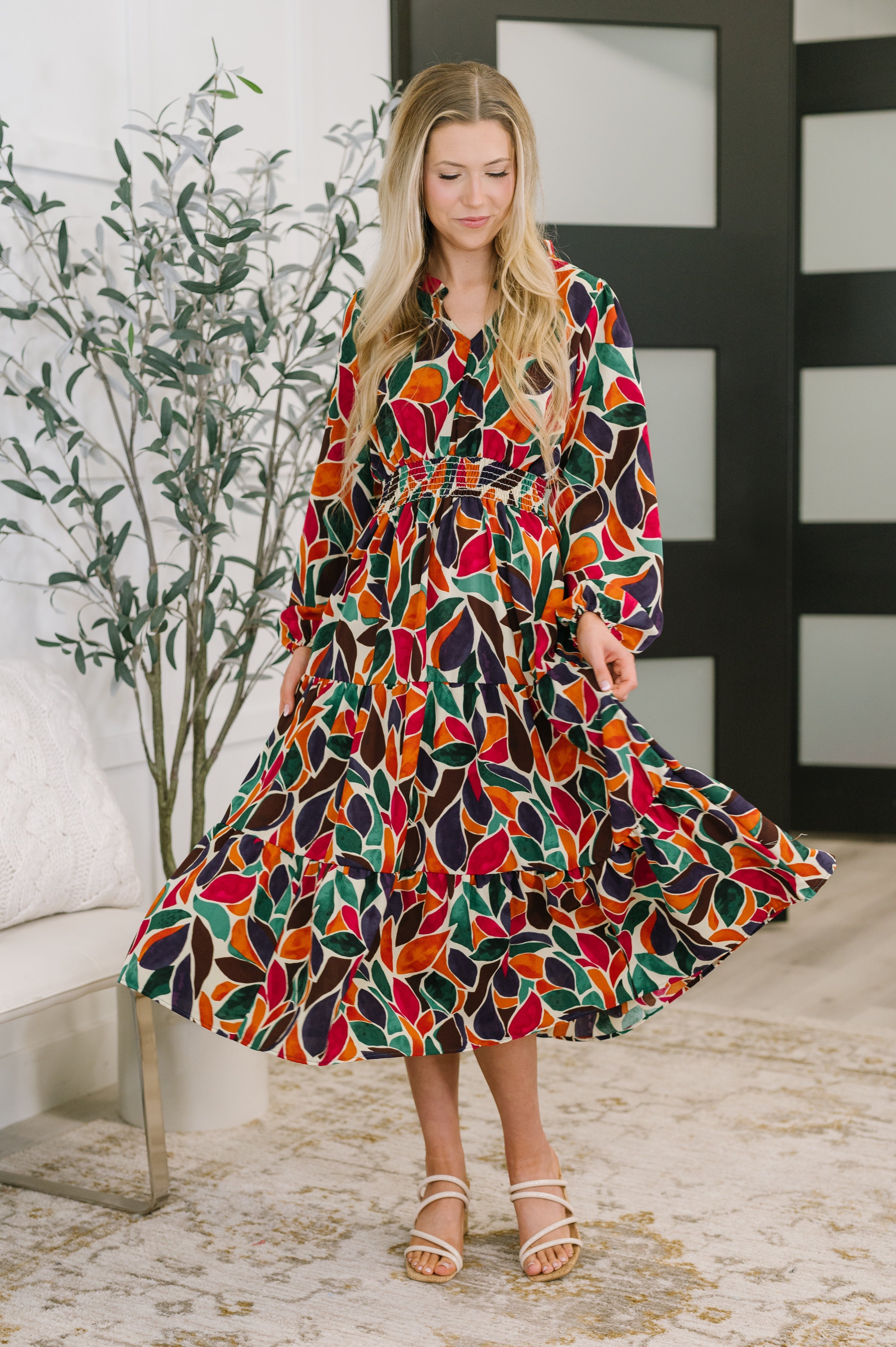 Woman wearing a long-sleeve tiered dress with colorful leaf patterns in an indoor setting with plants and modern decor