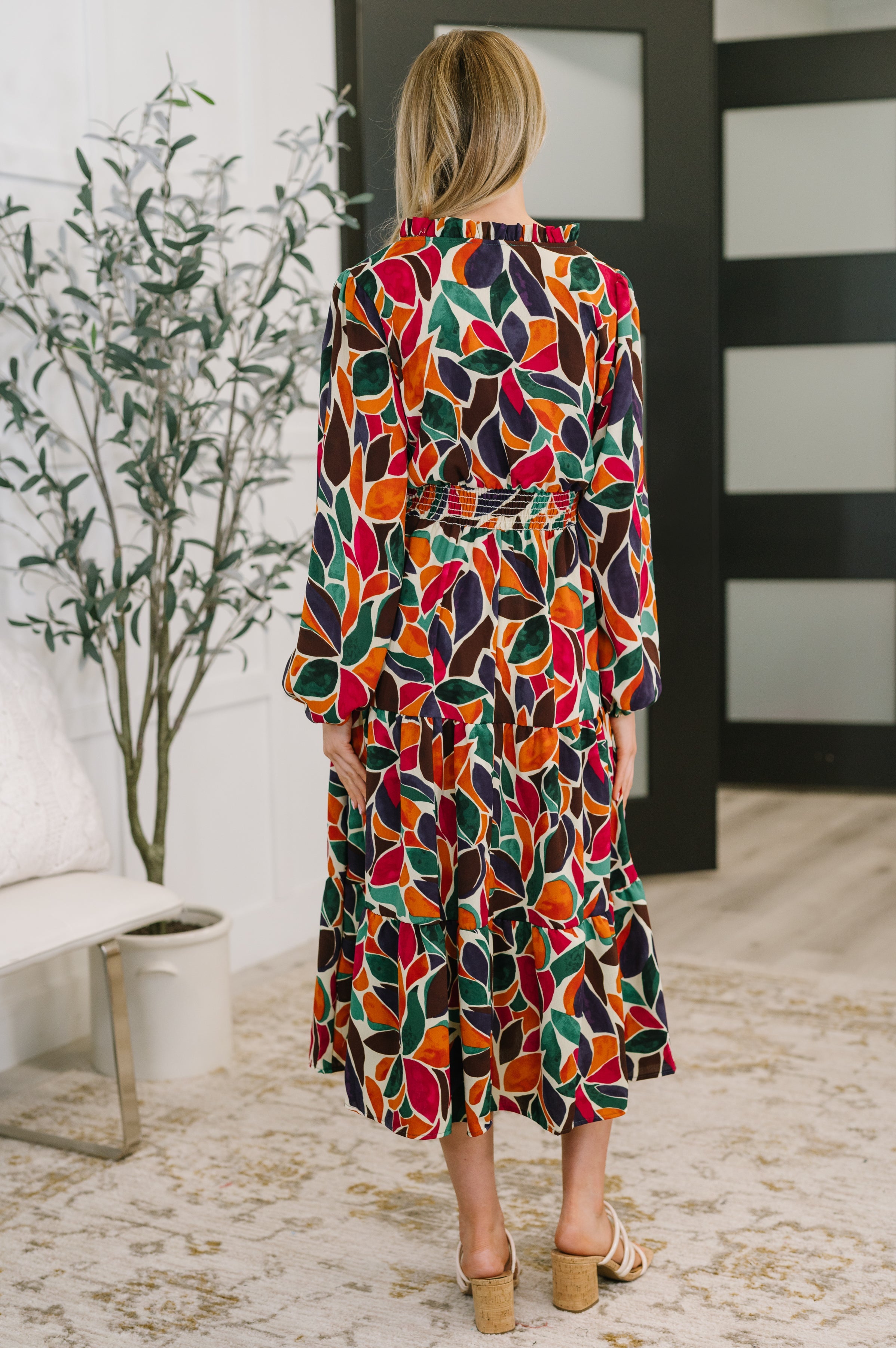 Woman wearing a colorful long sleeve tiered dress with bold abstract leaf shapes and a smocked waist standing indoors