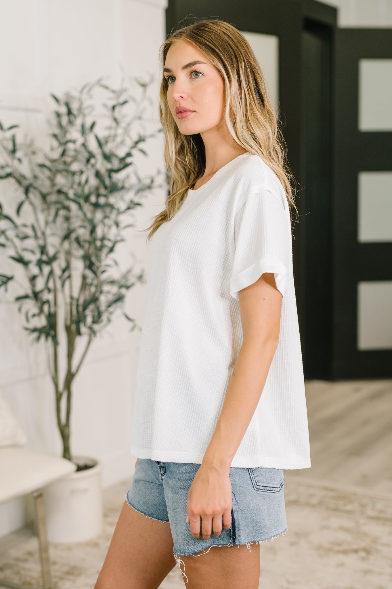 Side profile of woman in ivory waffle knit relaxed tee paired with light denim shorts in bright eclectic room