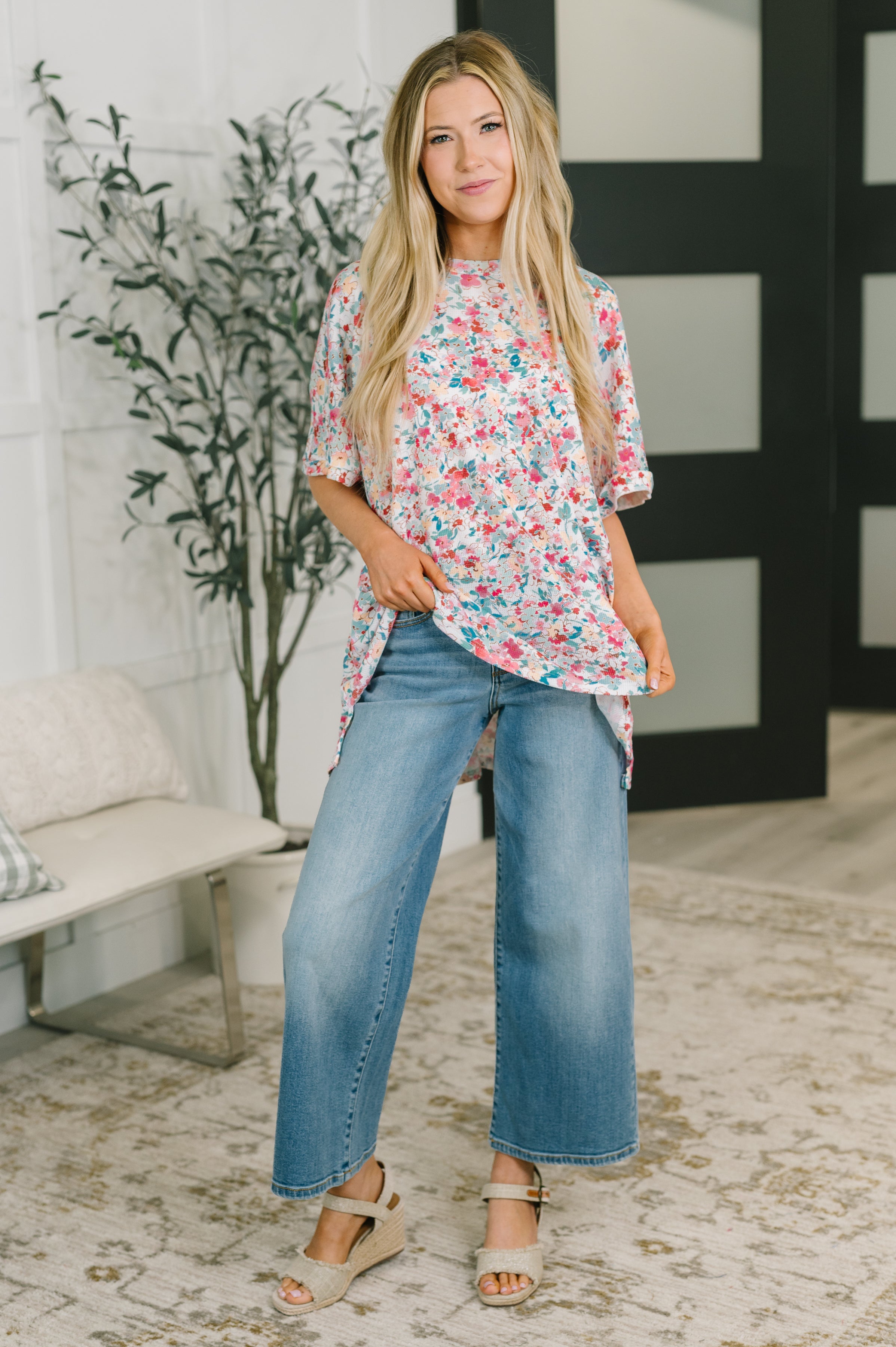 Woman wearing pink floral print dolman sleeve blouse with relaxed fit paired with wide-leg jeans and wedge sandals indoors