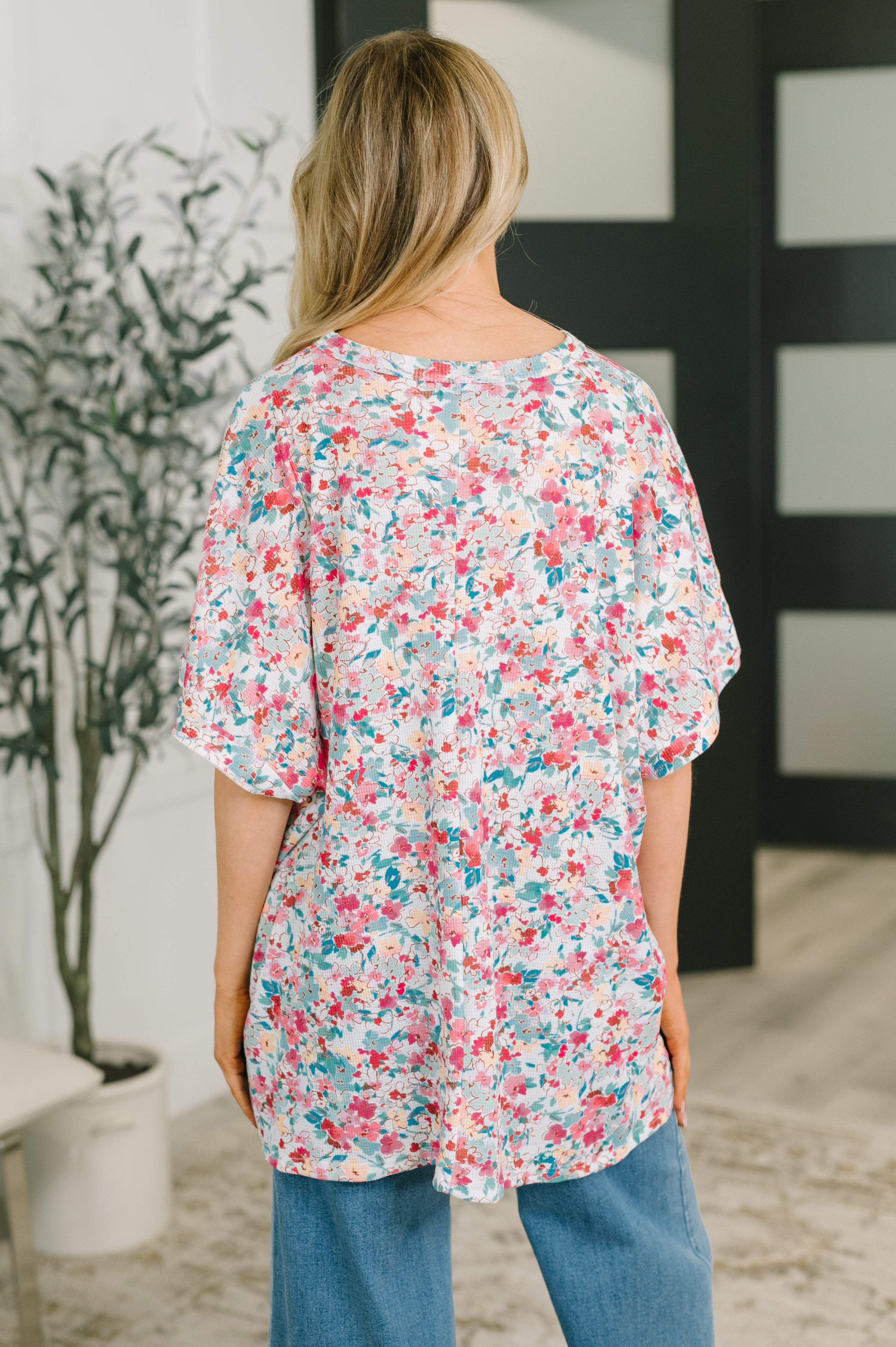 Back view of woman wearing flowy floral print blouse with dolman sleeves paired with loose blue jeans indoors