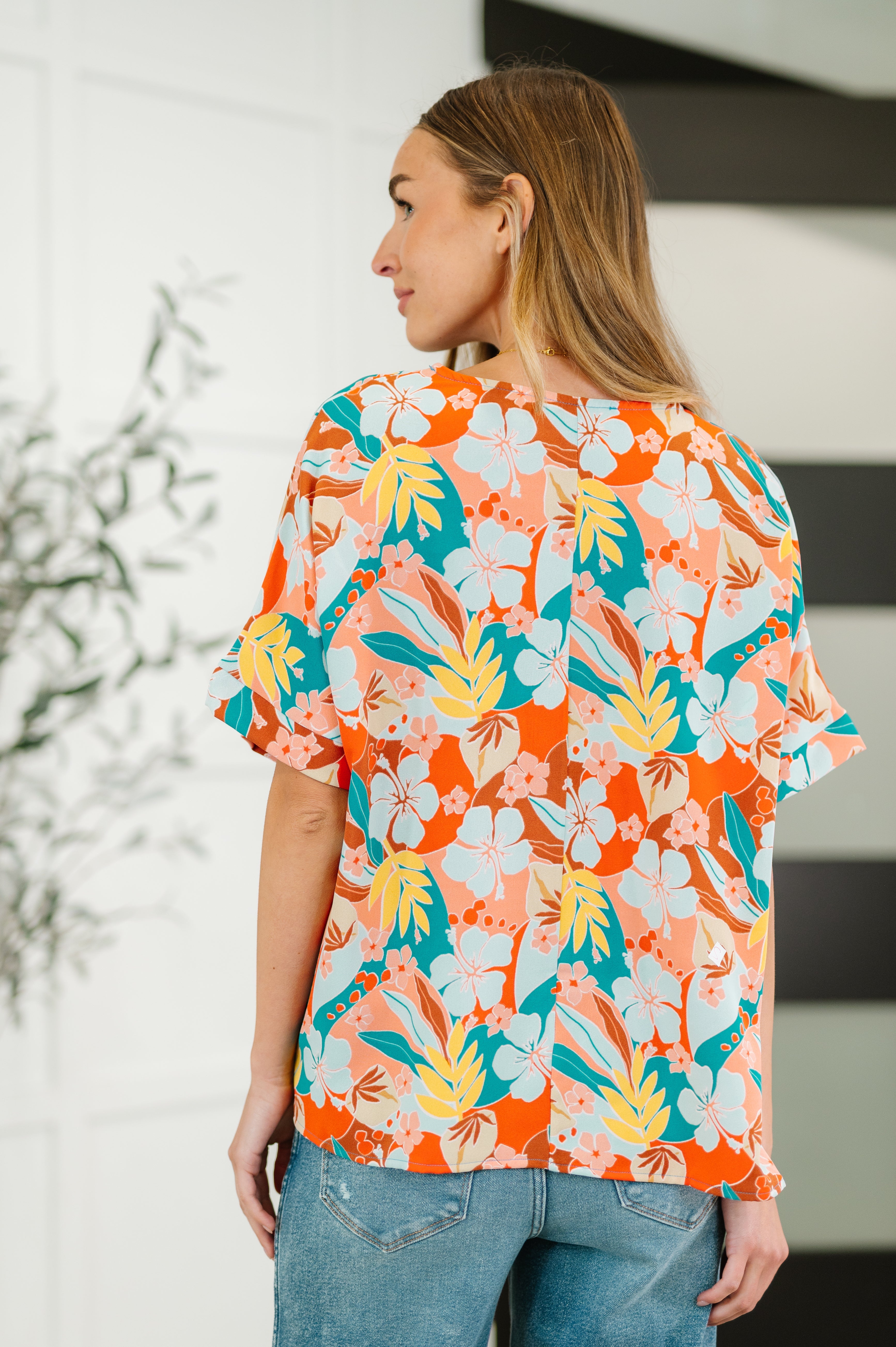 Back view of woman wearing vibrant orange floral print top with short sleeves paired with casual blue jeans indoors