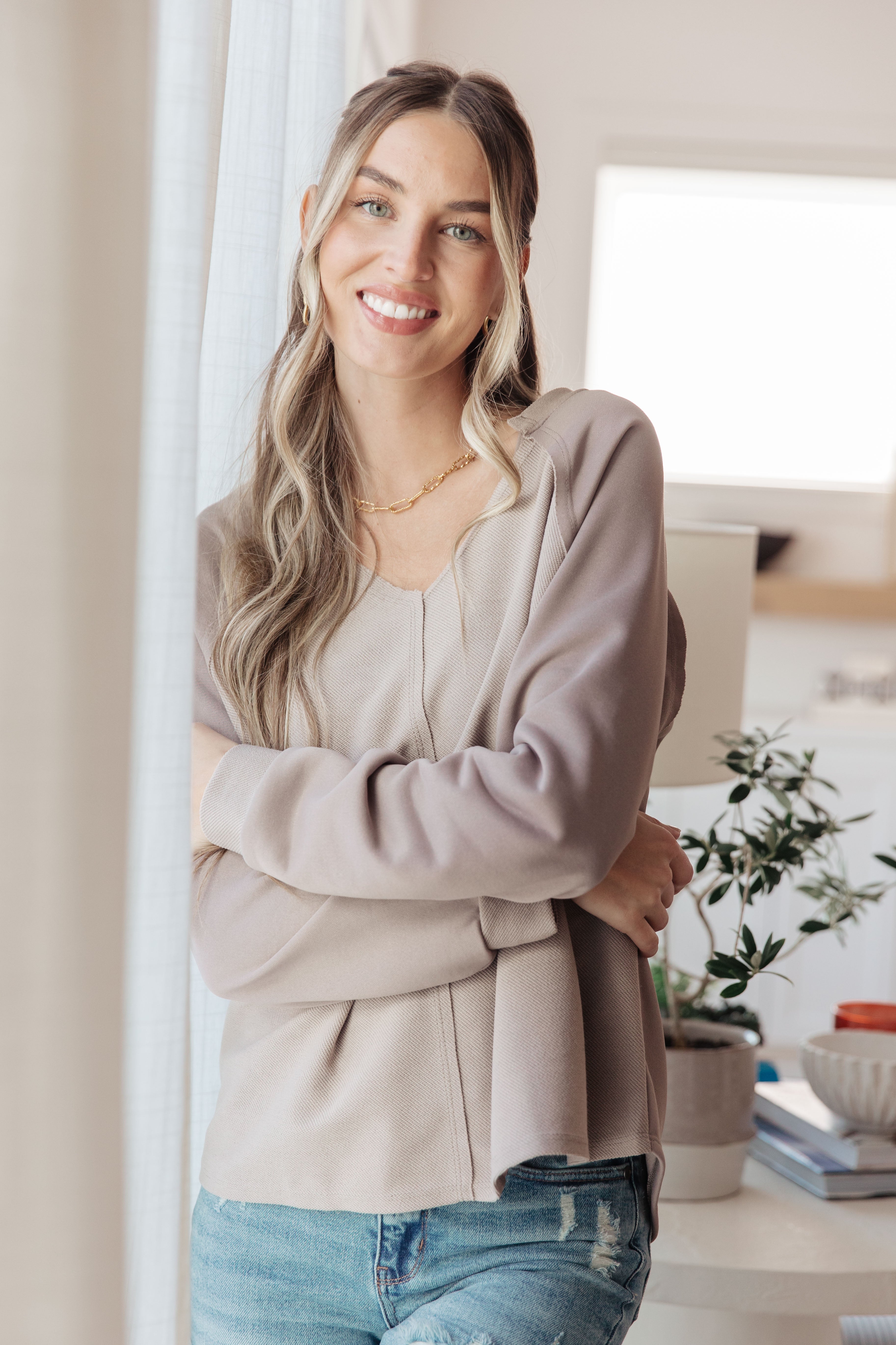 Smiling woman wearing a beige long sleeve top with a relaxed fit and V-neck posing indoors by a window