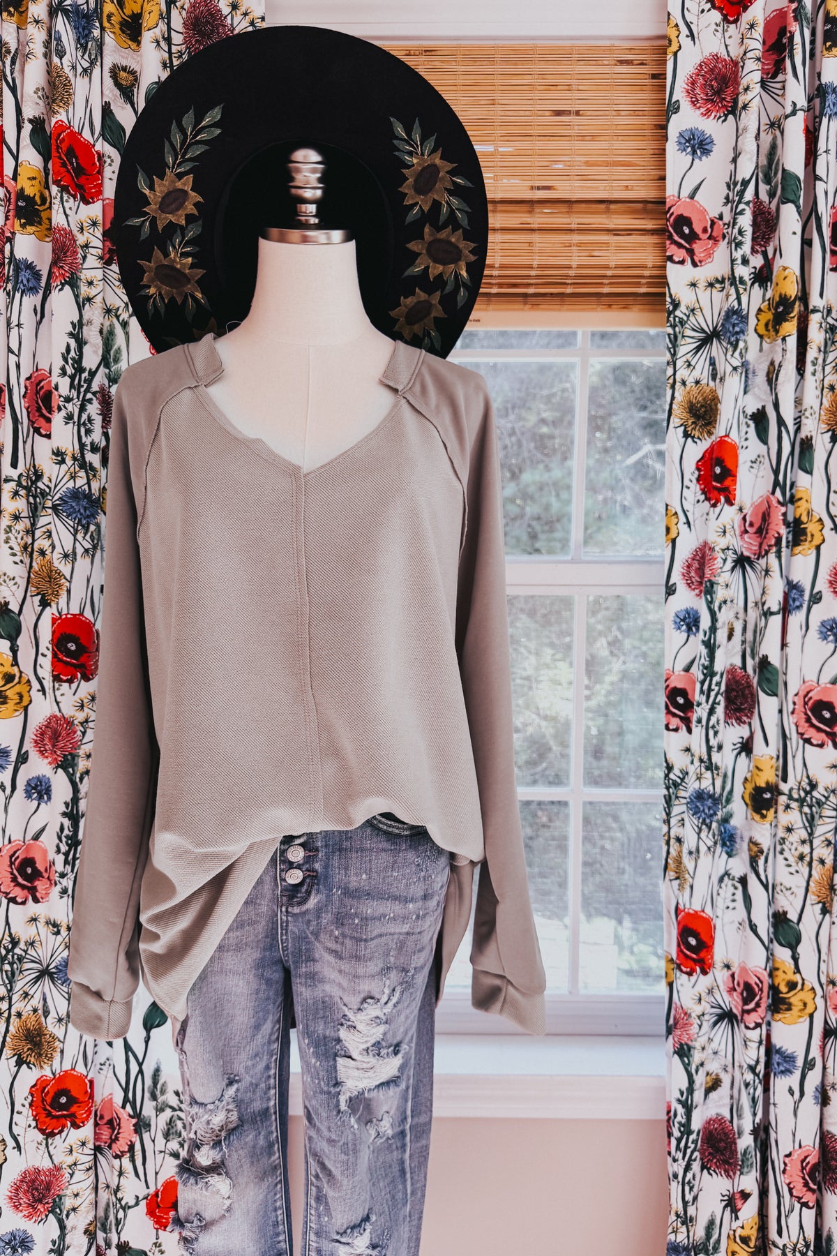 Beige textured knit top with a notched V neckline raglan sleeves and raw hem styled with distressed jeans on a mannequin by a window