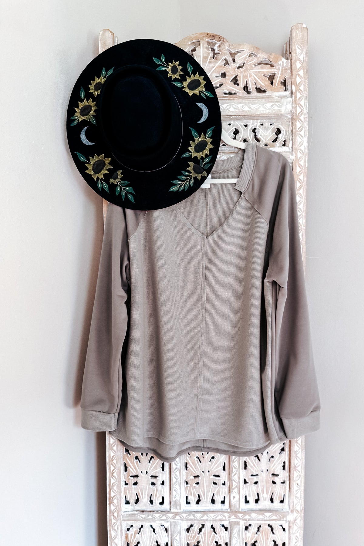 Beige textured knit top with a notched V-neckline and raglan sleeves hanging beneath a black floral embroidered hat on an ornate white rack