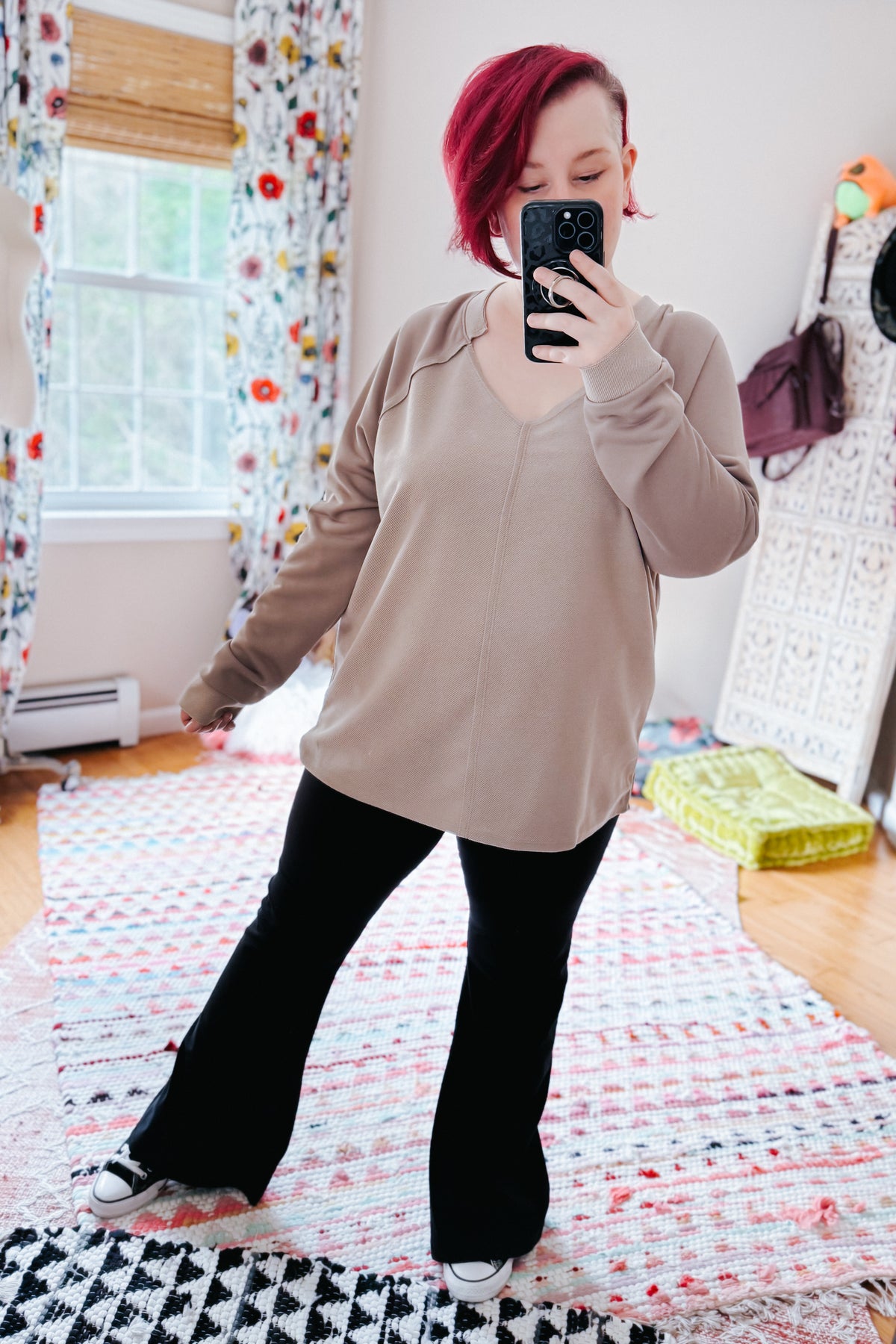 Woman taking a mirror selfie in a beige textured V neck knit top with raglan sleeves black flared pants and white sneakers in a bedroom