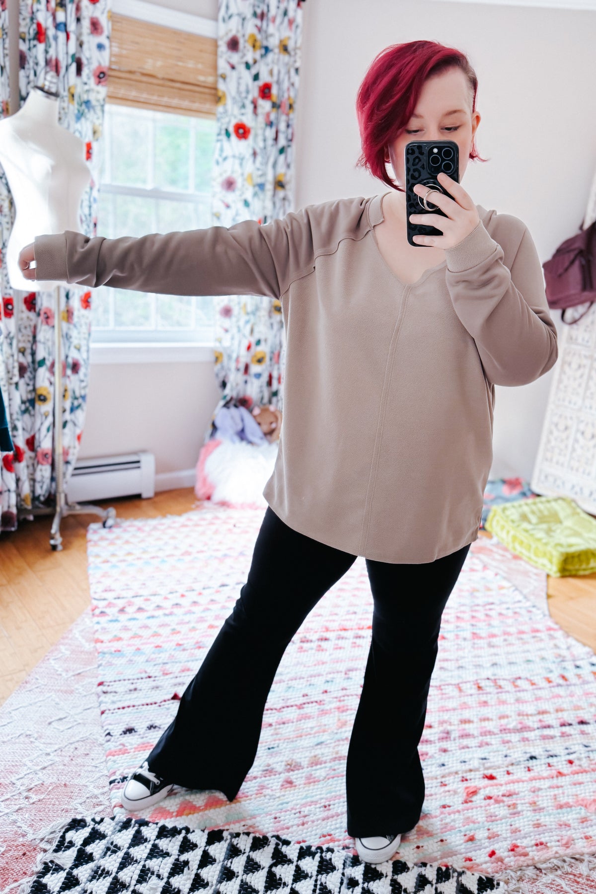 Woman taking a mirror selfie in a beige textured V-neck top with raglan sleeves, black flared pants, and sneakers in a colorful bedroom