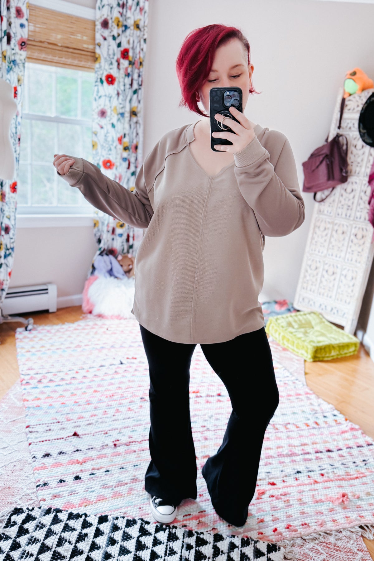 Red haired woman taking a mirror selfie in a beige textured V neck top with raglan sleeves black flared pants and sneakers in a colorful bedroom