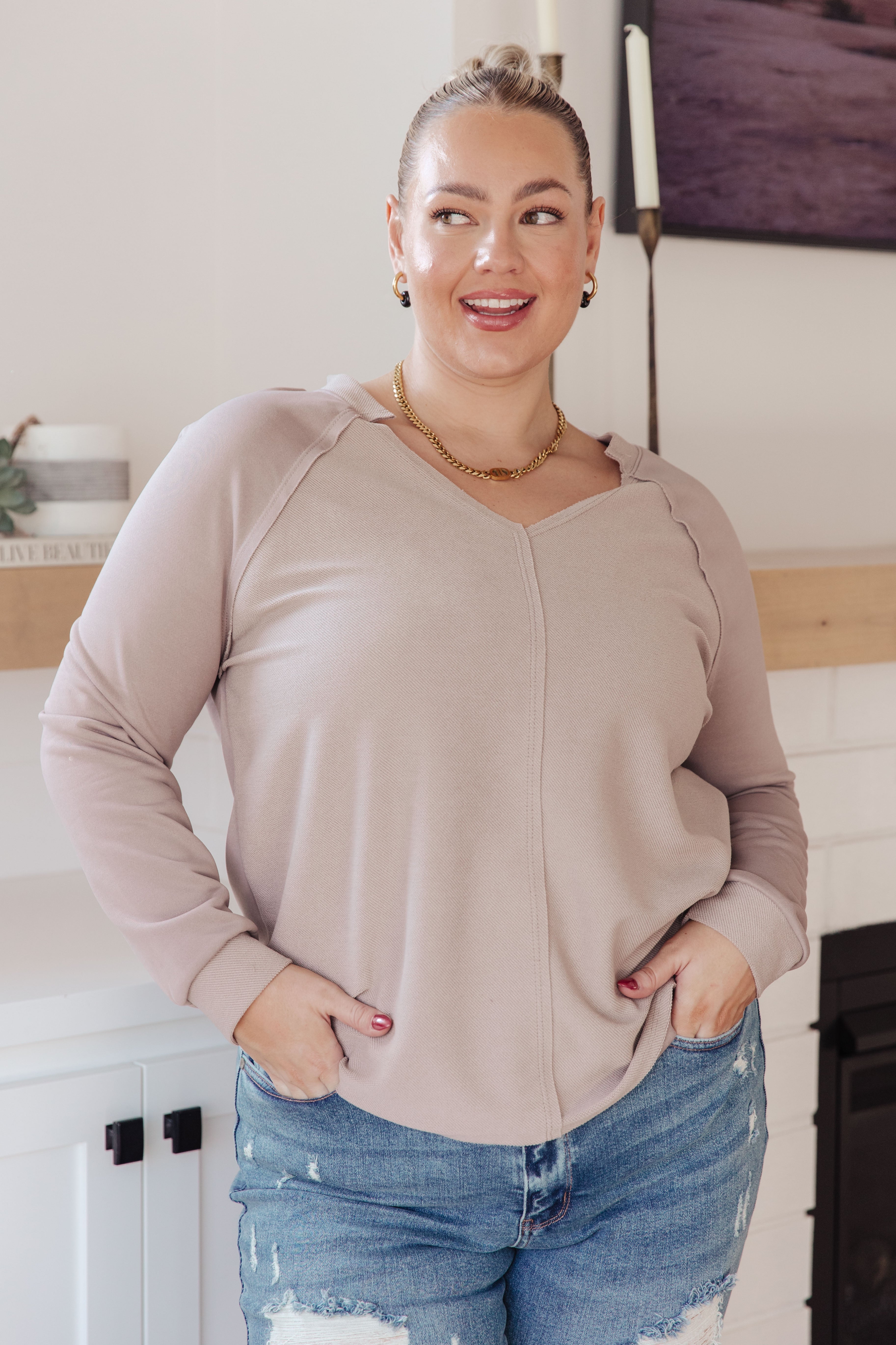 Woman smiling wearing a beige V-neck long sleeve top with relaxed fit and distressed blue jeans indoors