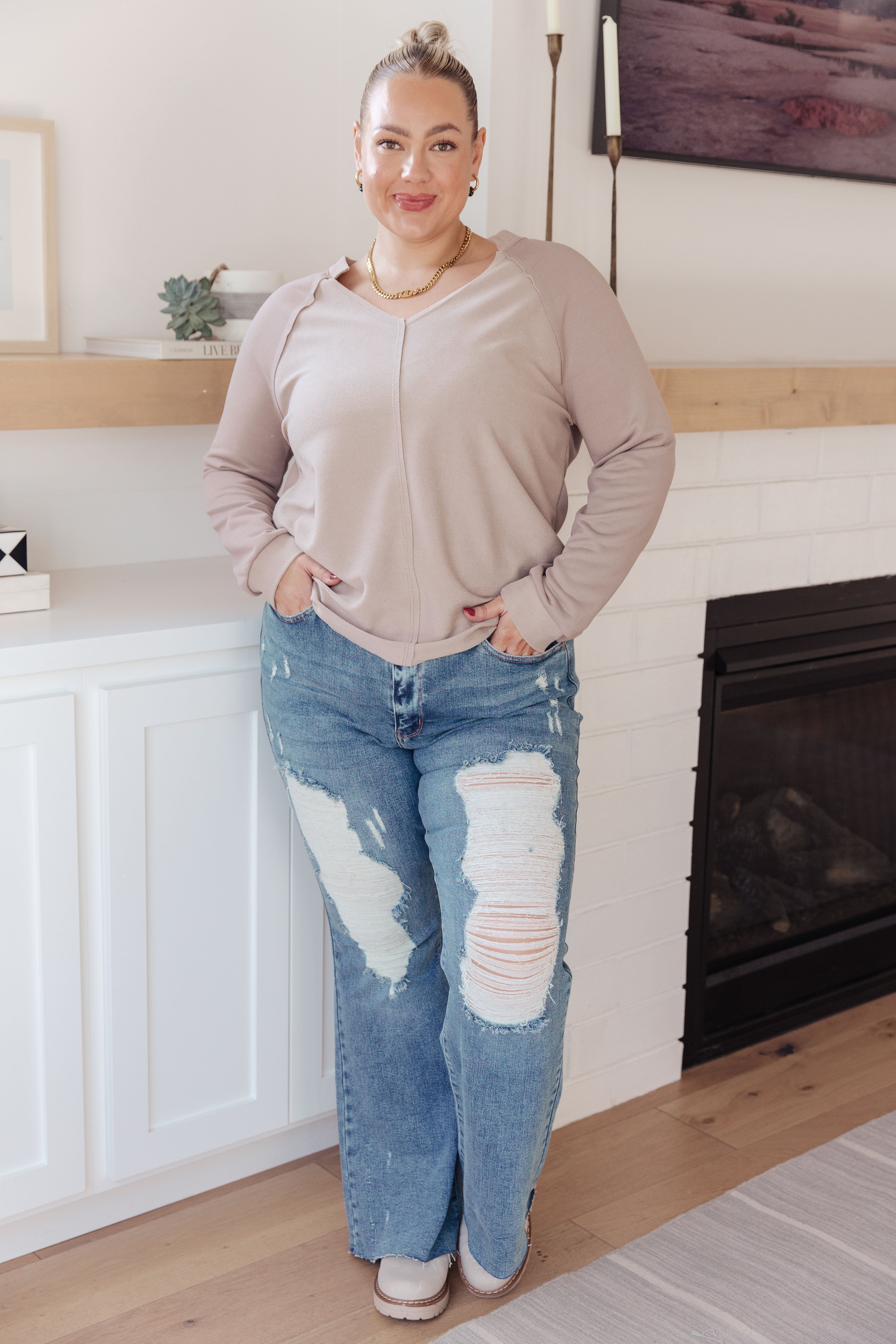 Woman standing indoors wearing a beige V-neck long sleeve top paired with distressed blue jeans and neutral shoes