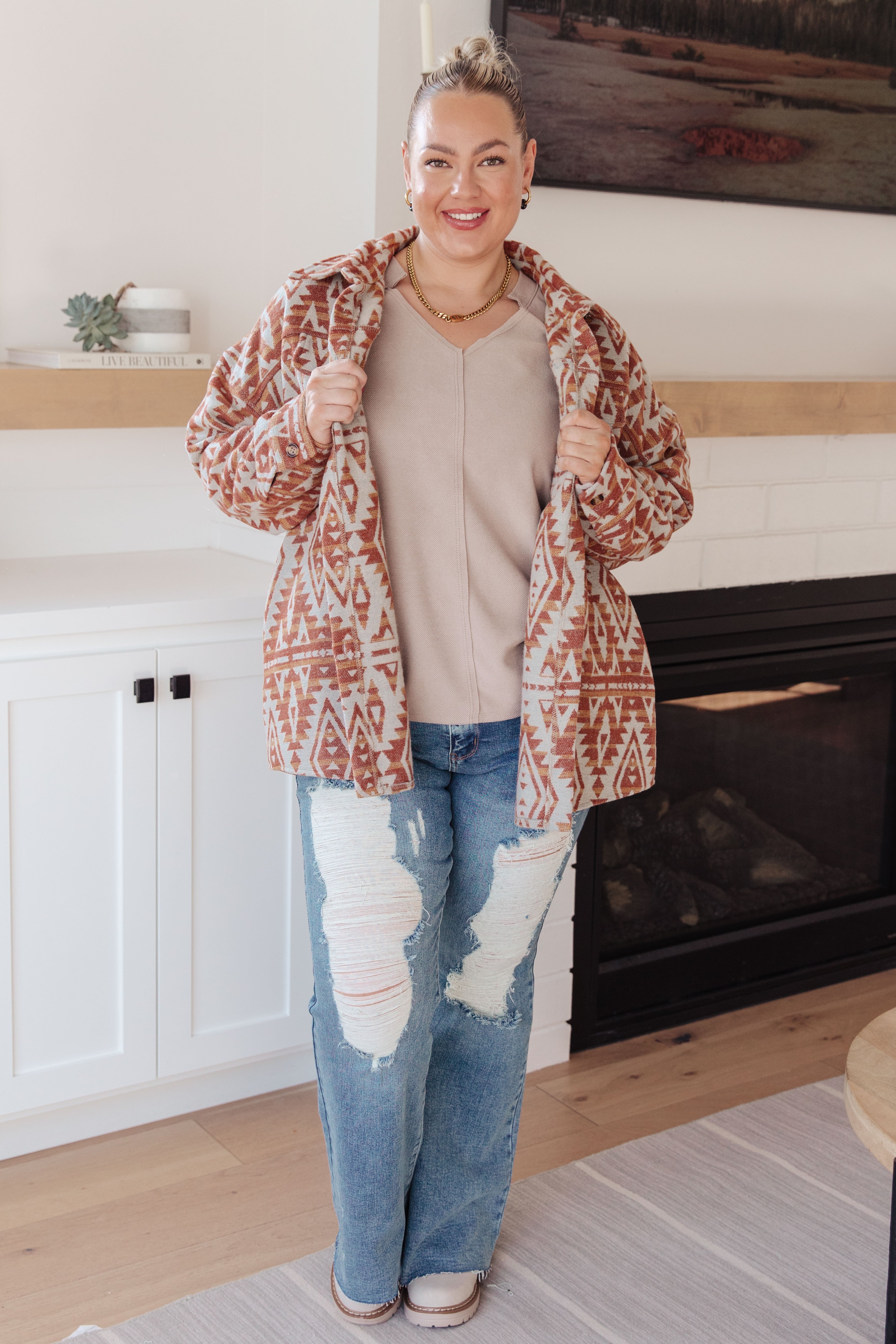 Smiling woman indoors wearing a beige top with a V-neck and an oversized patterned jacket with distressed blue jeans