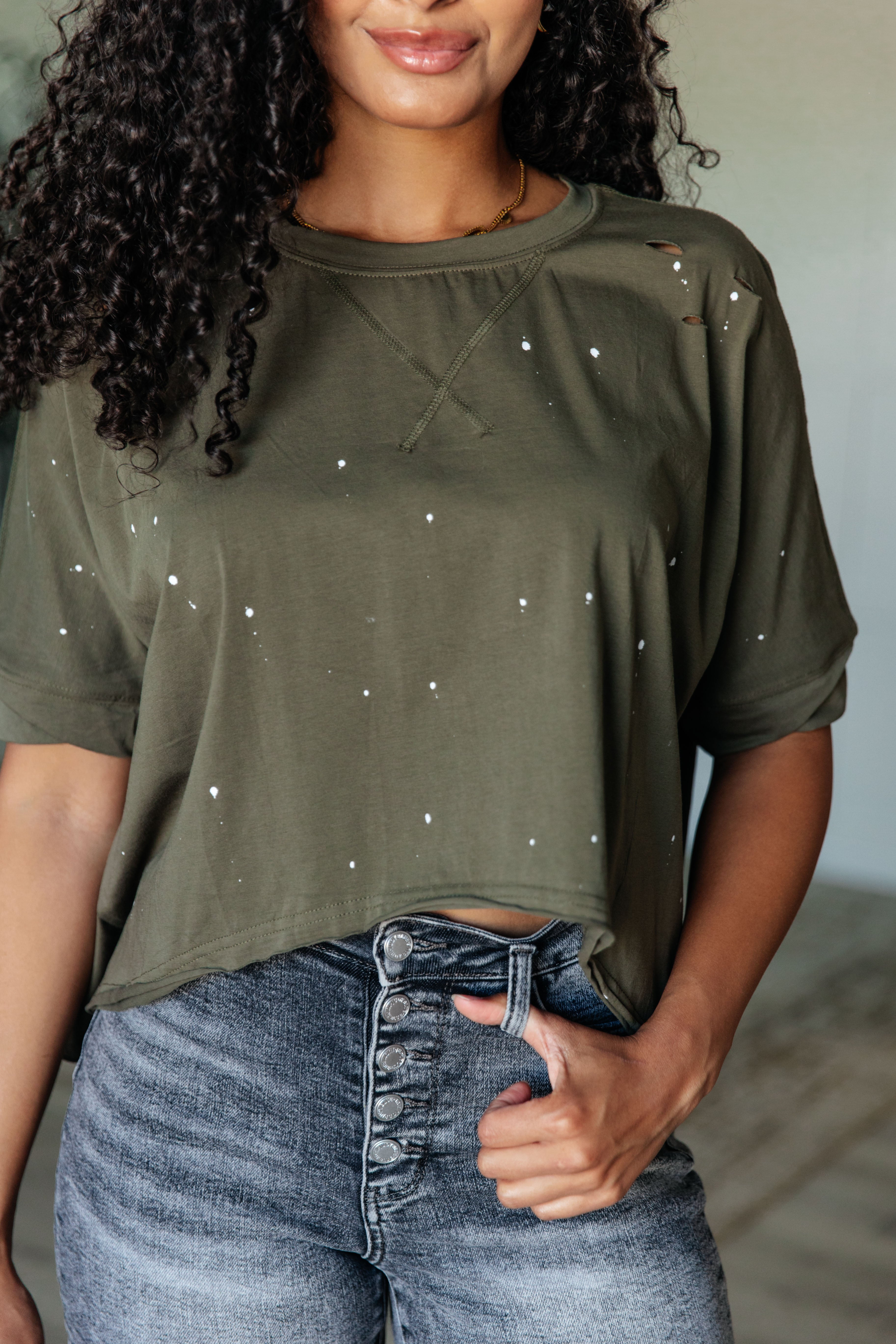 Woman with curly hair wearing an olive distressed cropped short sleeve top paired with high-rise button fly jeans indoors