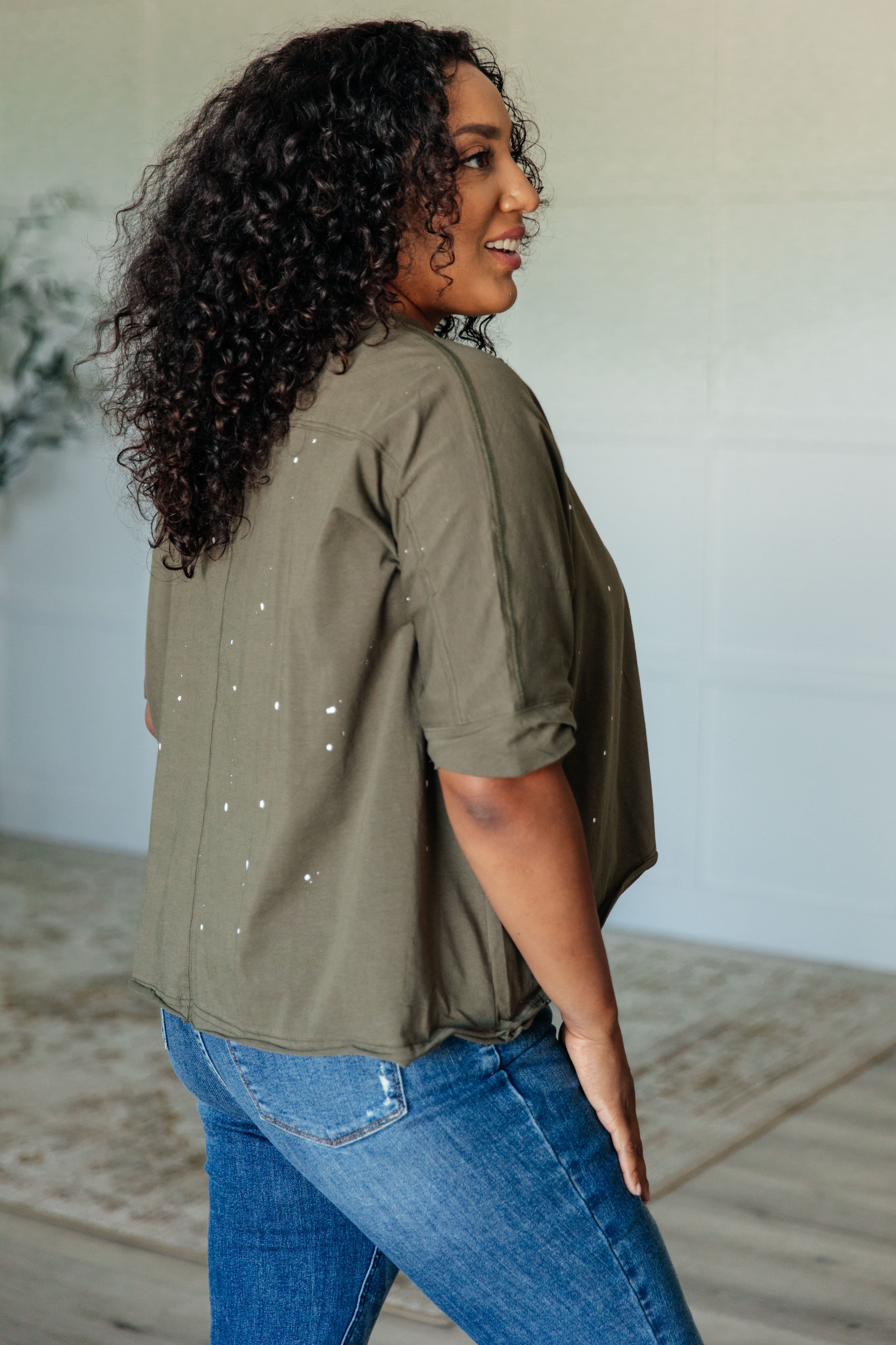 Woman with curly hair wearing an olive short sleeve top with subtle paint splatter details and blue jeans indoors