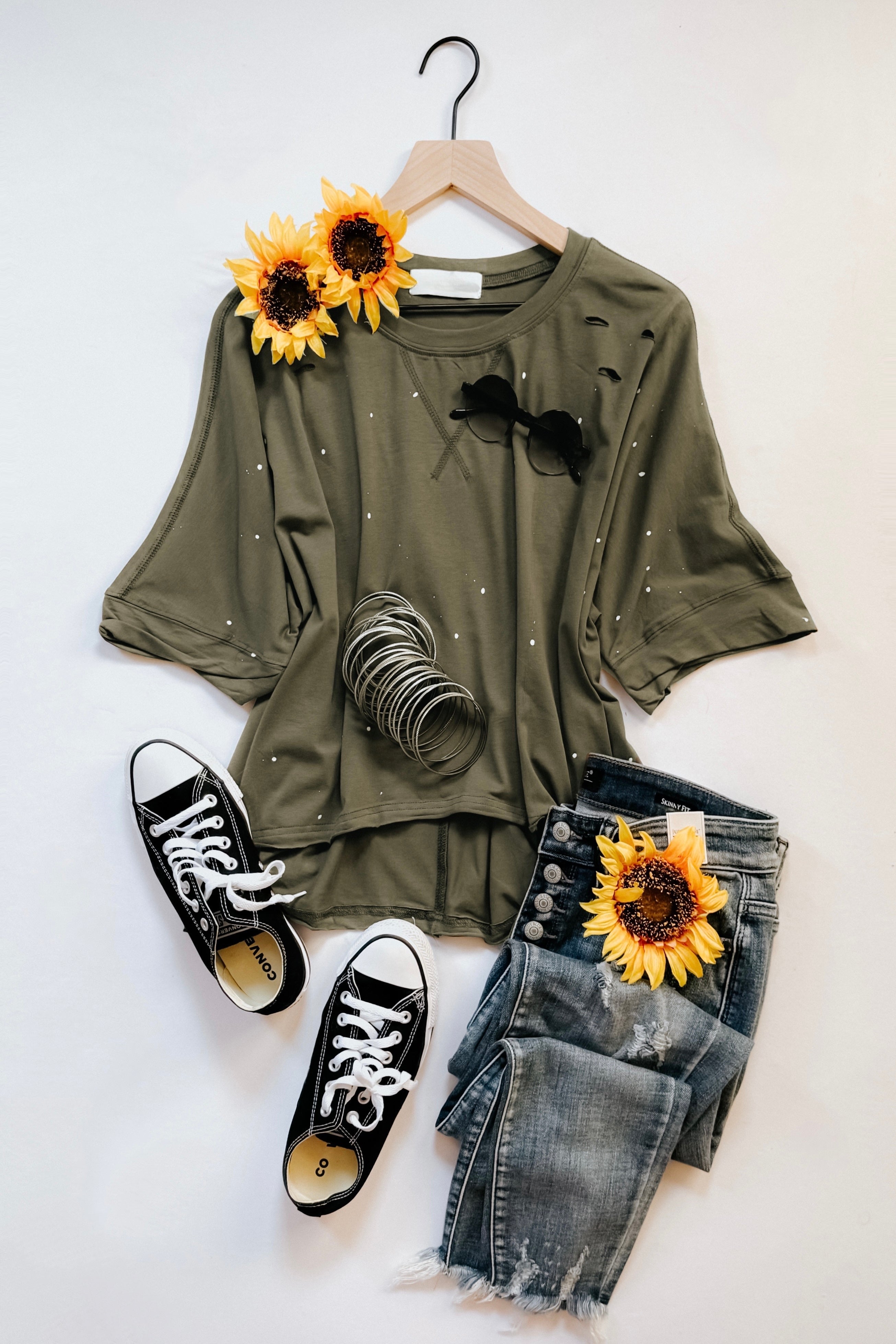 Olive relaxed fit short sleeve top with small white paint speckles displayed with distressed blue jeans and black sneakers