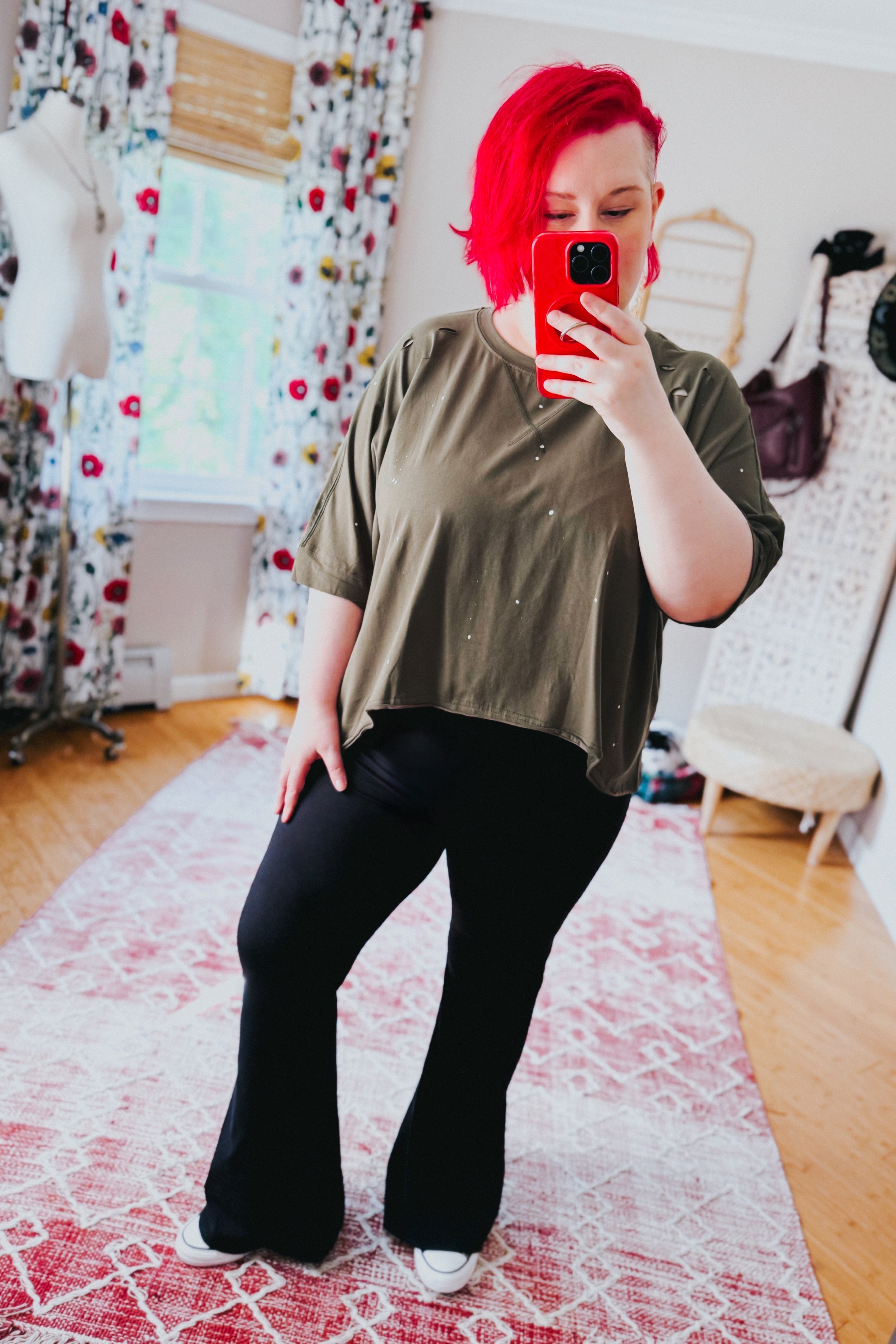 Woman with bright red hair wearing an olive oversized short sleeve top with paint splatter and black flared pants indoors