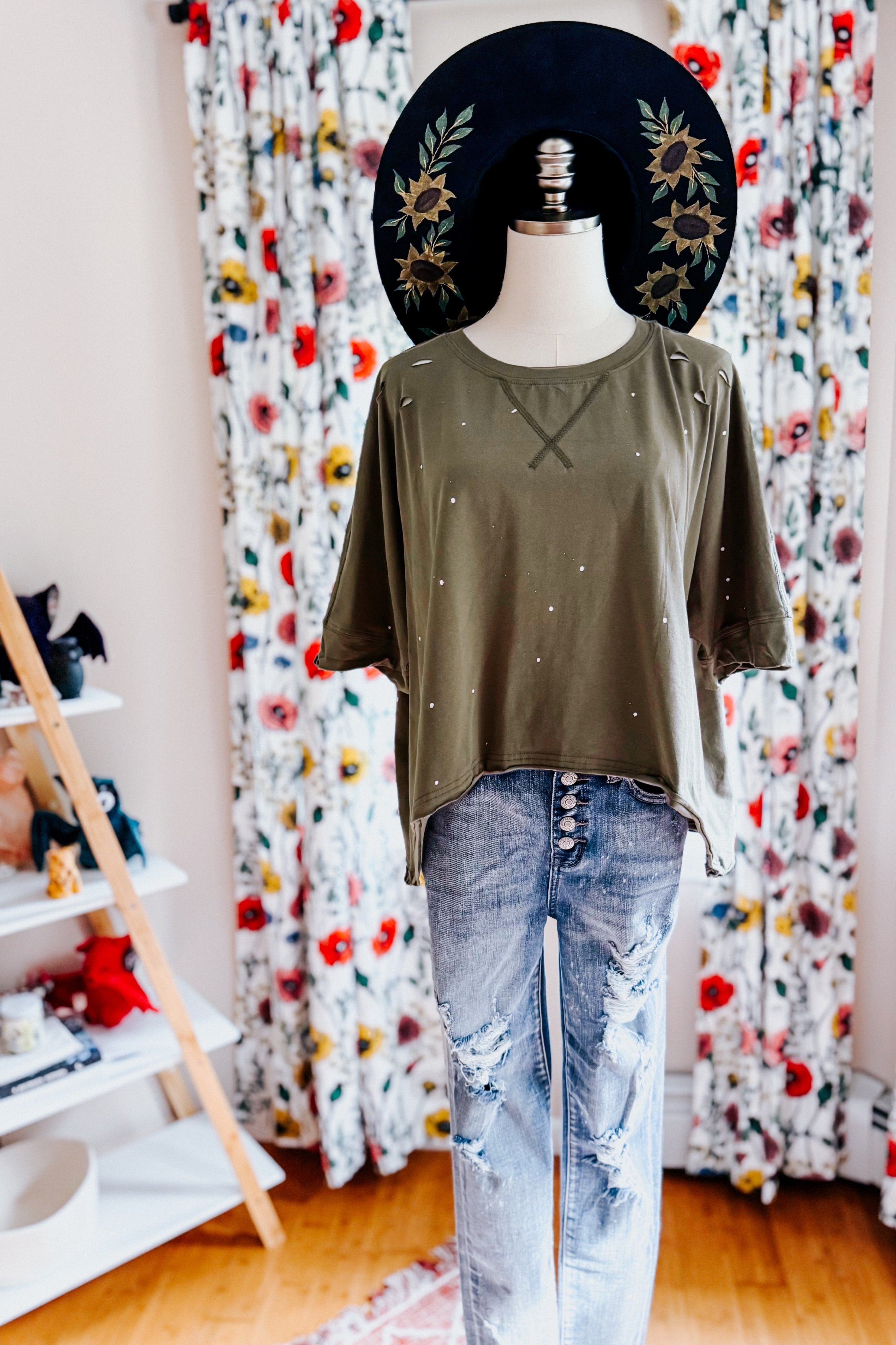 Mannequin wearing an olive loose fit short sleeve top with subtle paint splatter details and distressed light wash jeans indoors