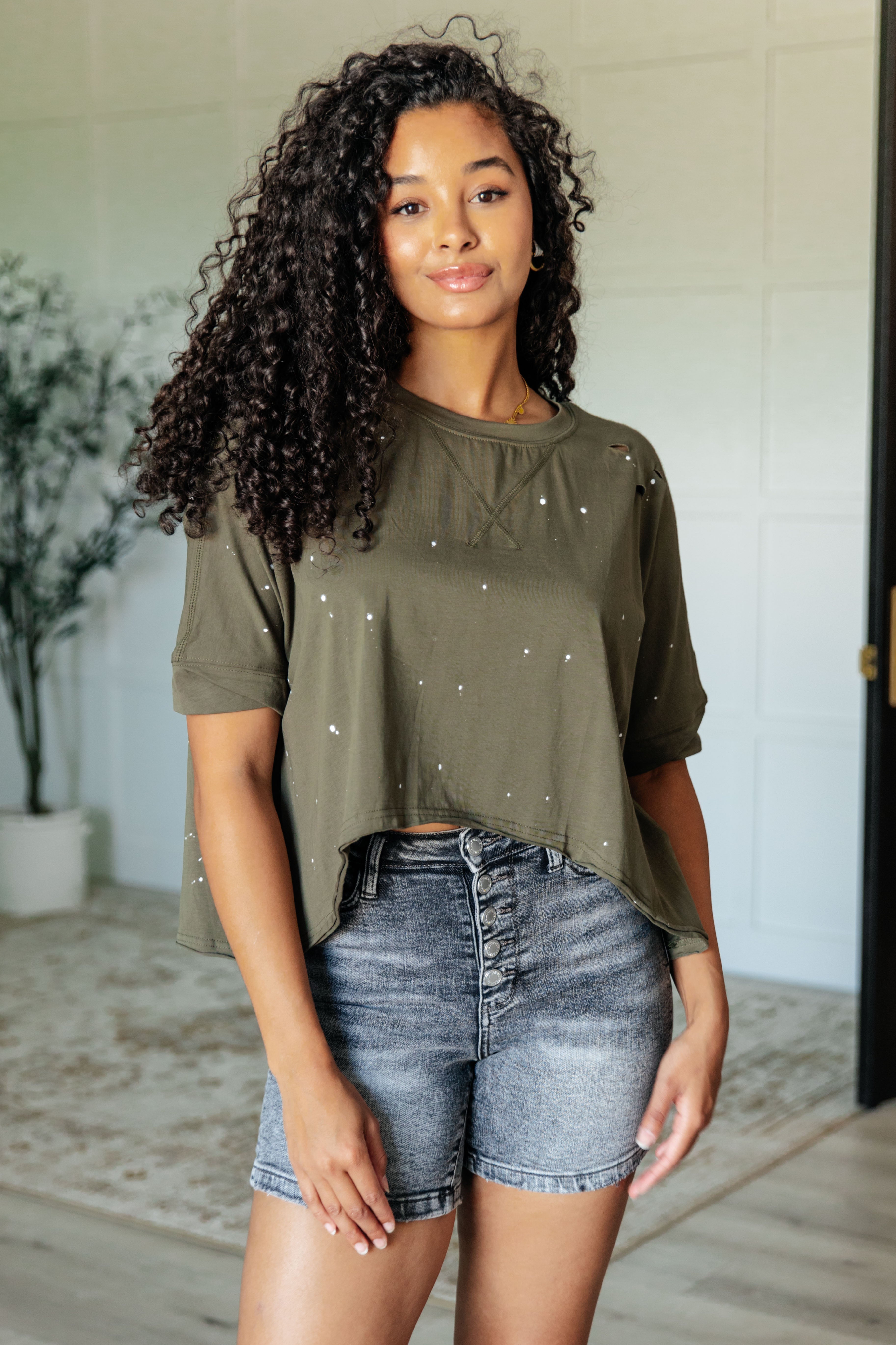 Woman with curly hair wearing an olive asymmetrical distressed short sleeve top and high-waisted denim shorts indoors