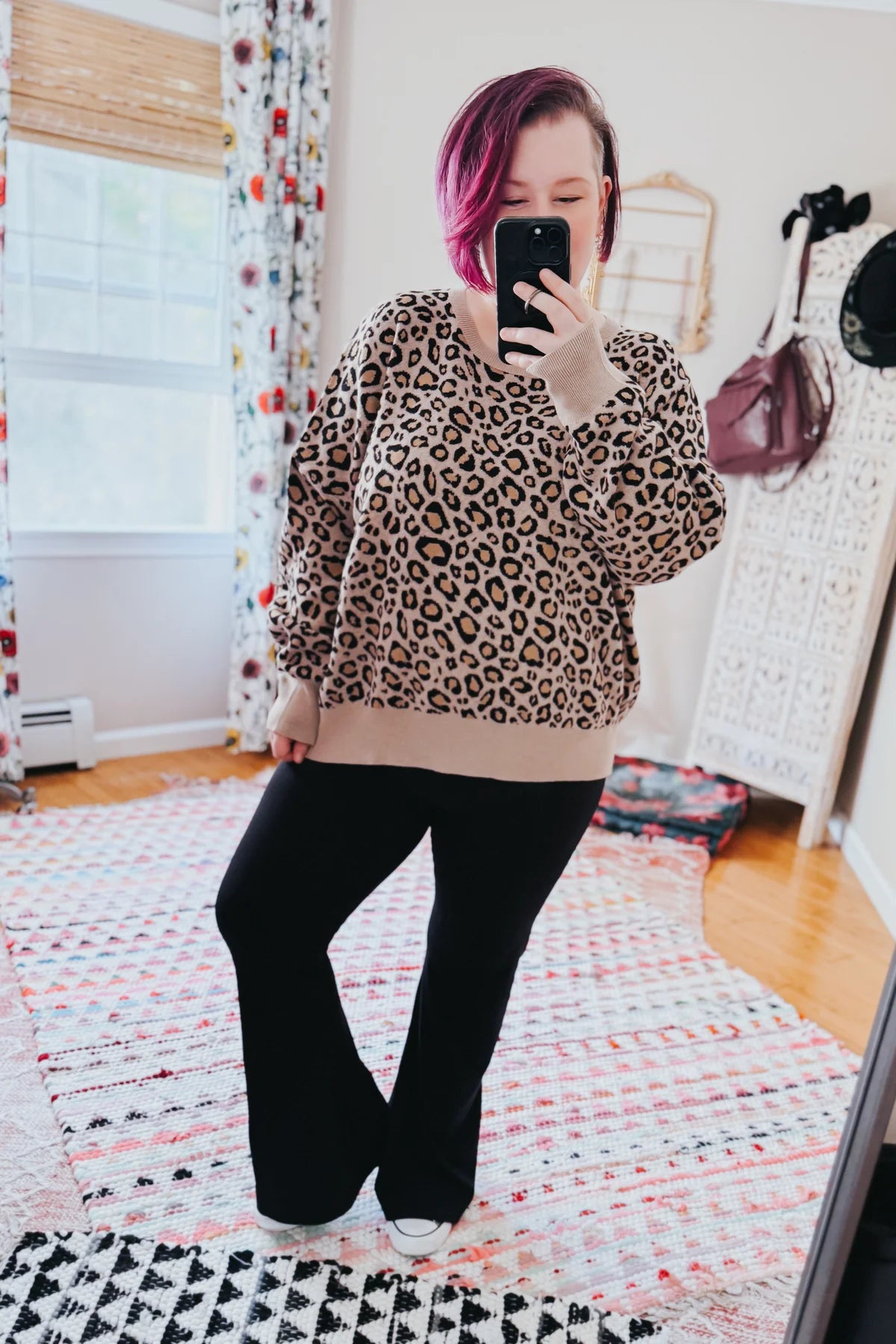 Woman taking a mirror selfie wearing a loose brown leopard print top with beige cuffs and black pants indoors