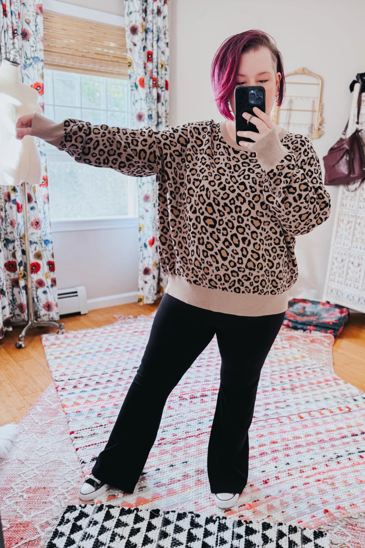 Woman taking a mirror selfie wearing a brown leopard print pullover with beige cuffs and black pants indoors