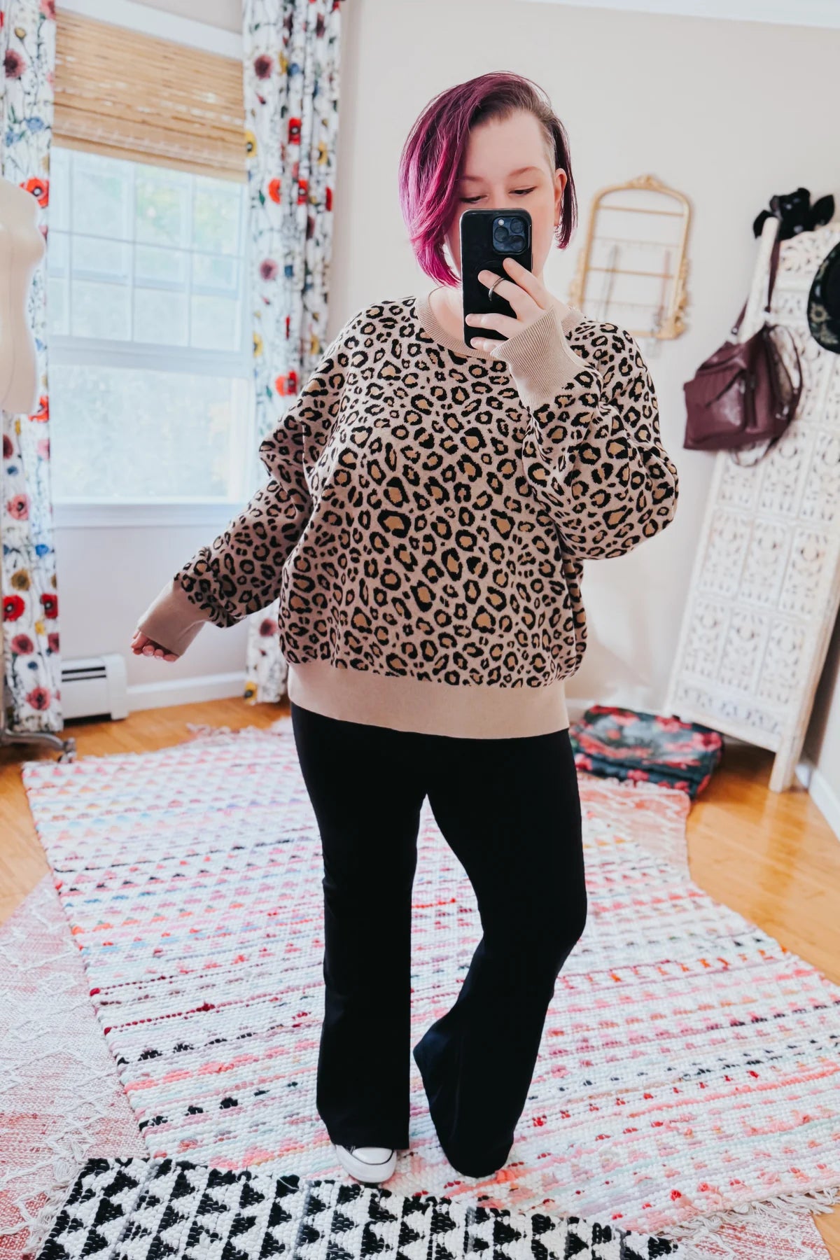 Woman taking a mirror selfie wearing a brown leopard print pullover with beige cuffs and black pants indoors