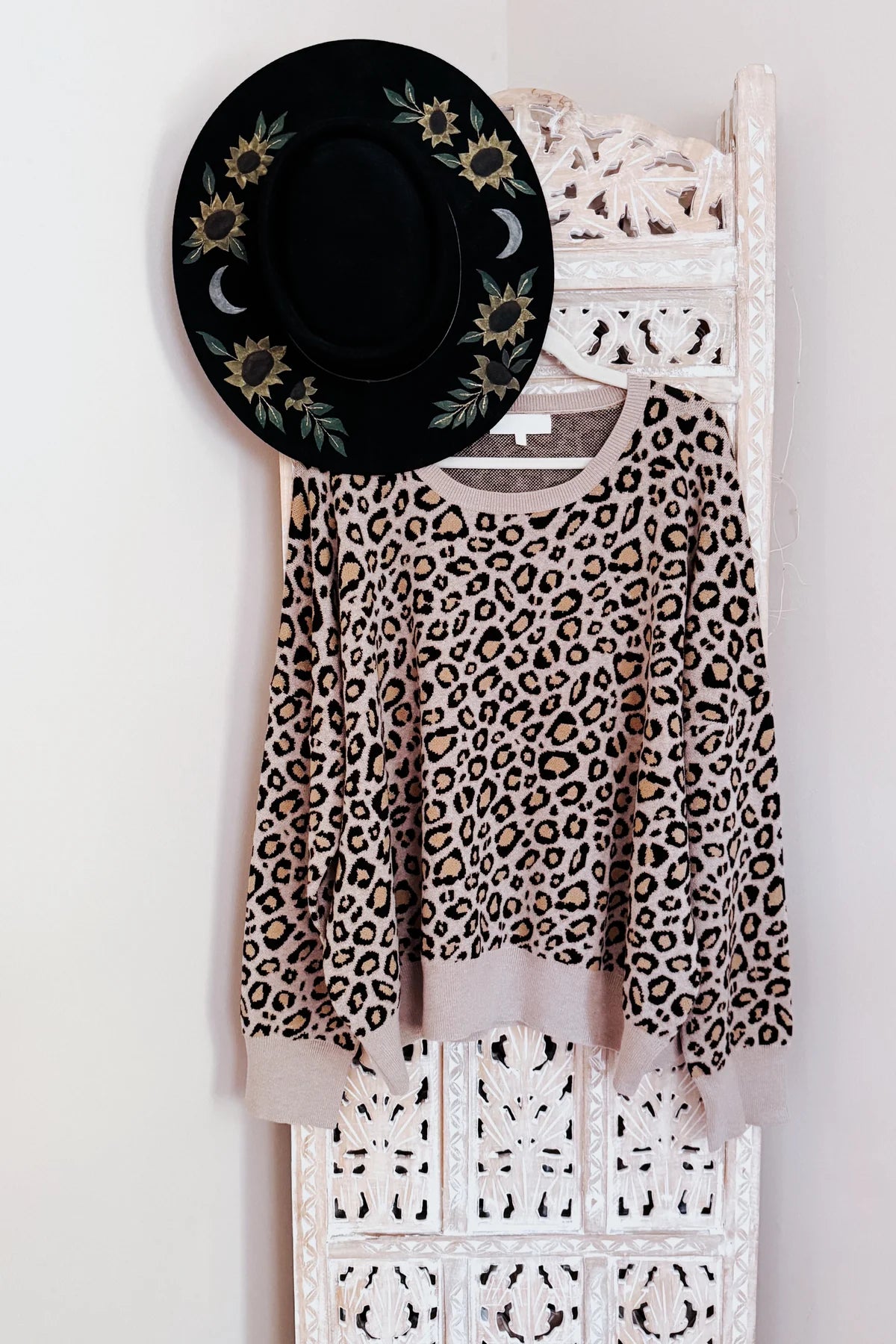 Brown leopard print long sleeve sweater hanging on carved white wooden panel with black floral hat above