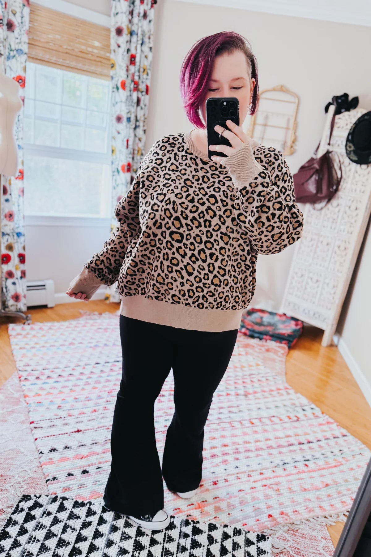 Woman taking a mirror selfie wearing a brown leopard print sweater with beige cuffs and black pants indoors
