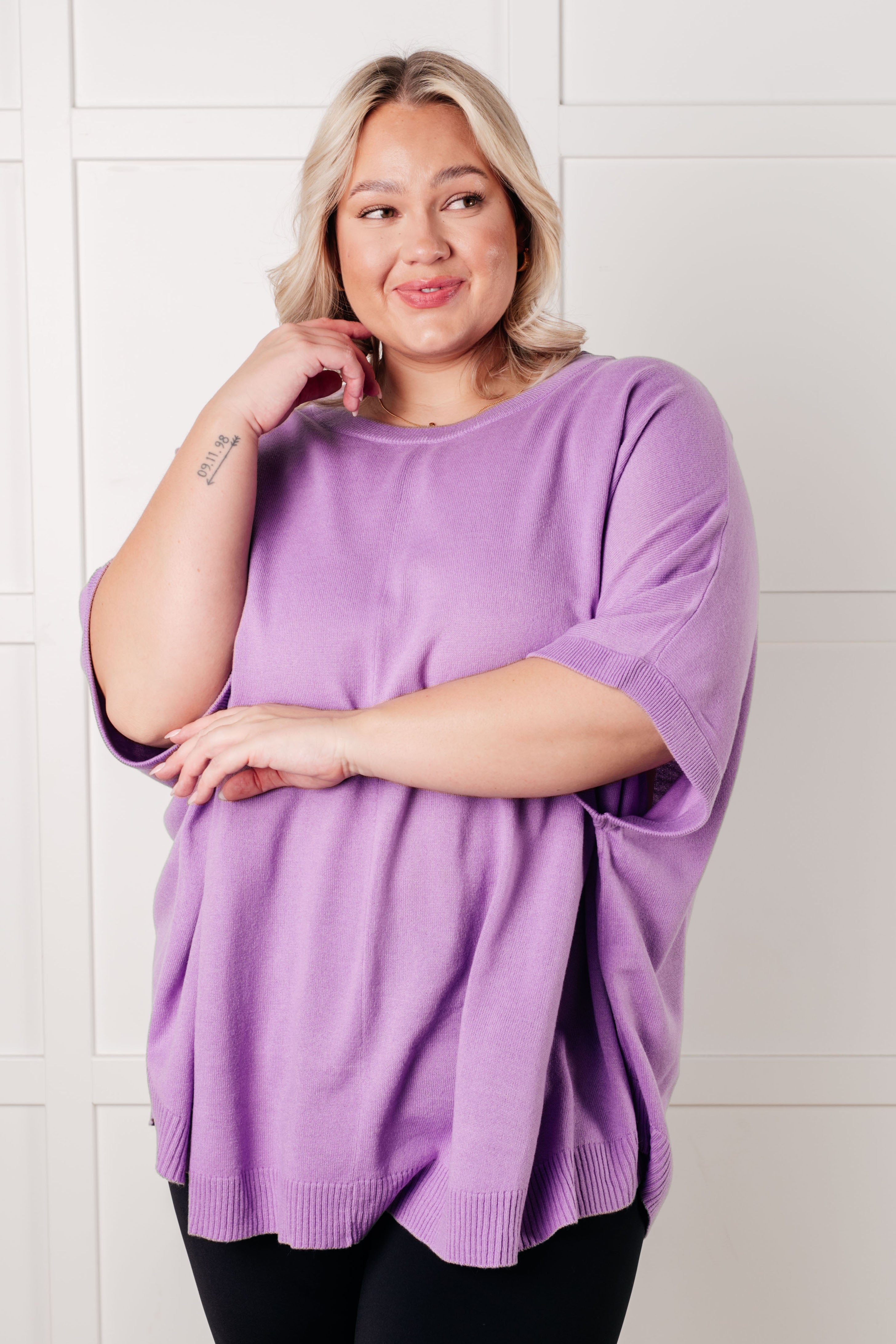 Smiling woman wearing a loose lilac dolman sleeve top paired with black pants against a white background