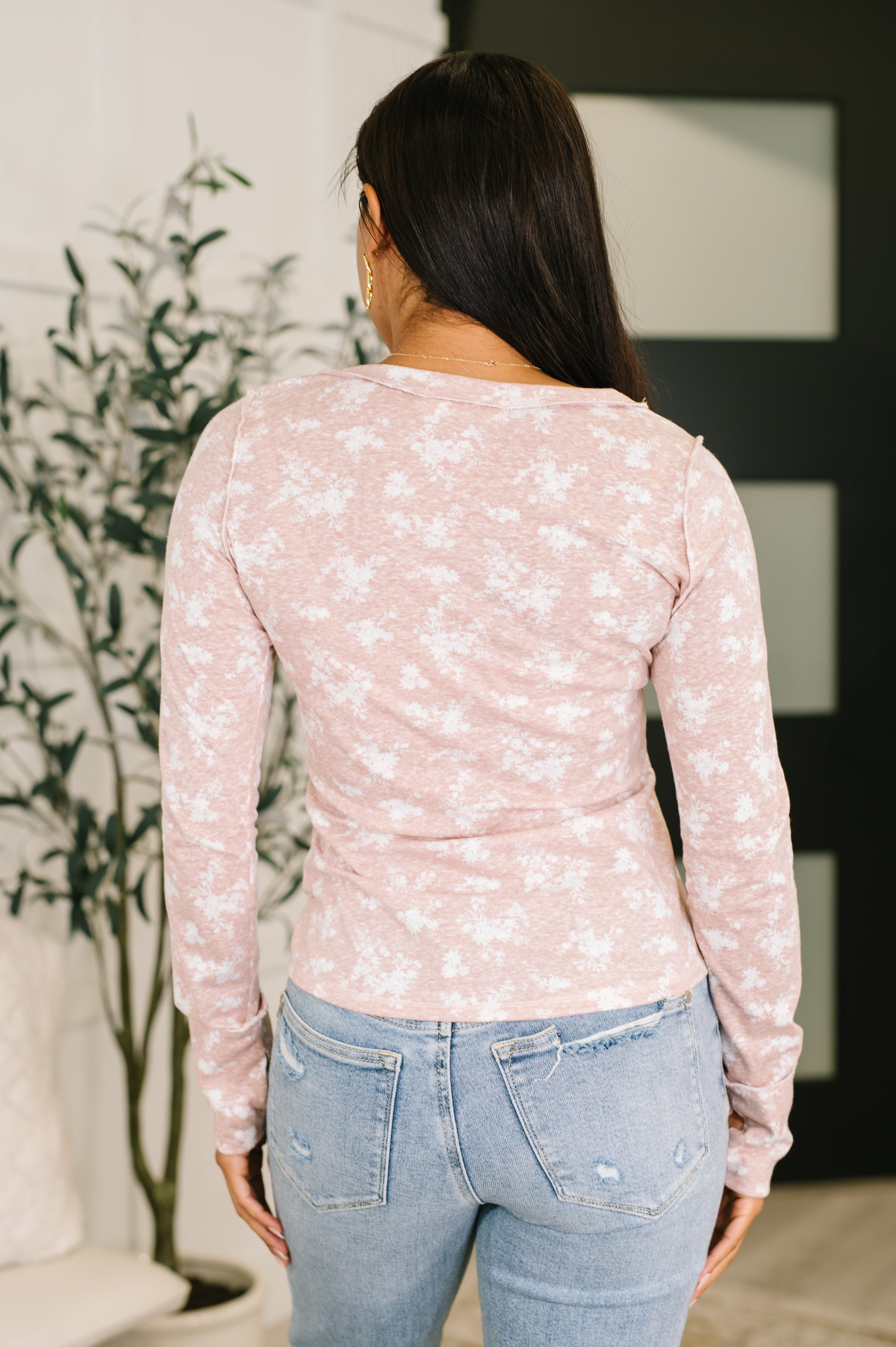 Back view of woman wearing a mauve long sleeve top with white floral print and light blue jeans indoors