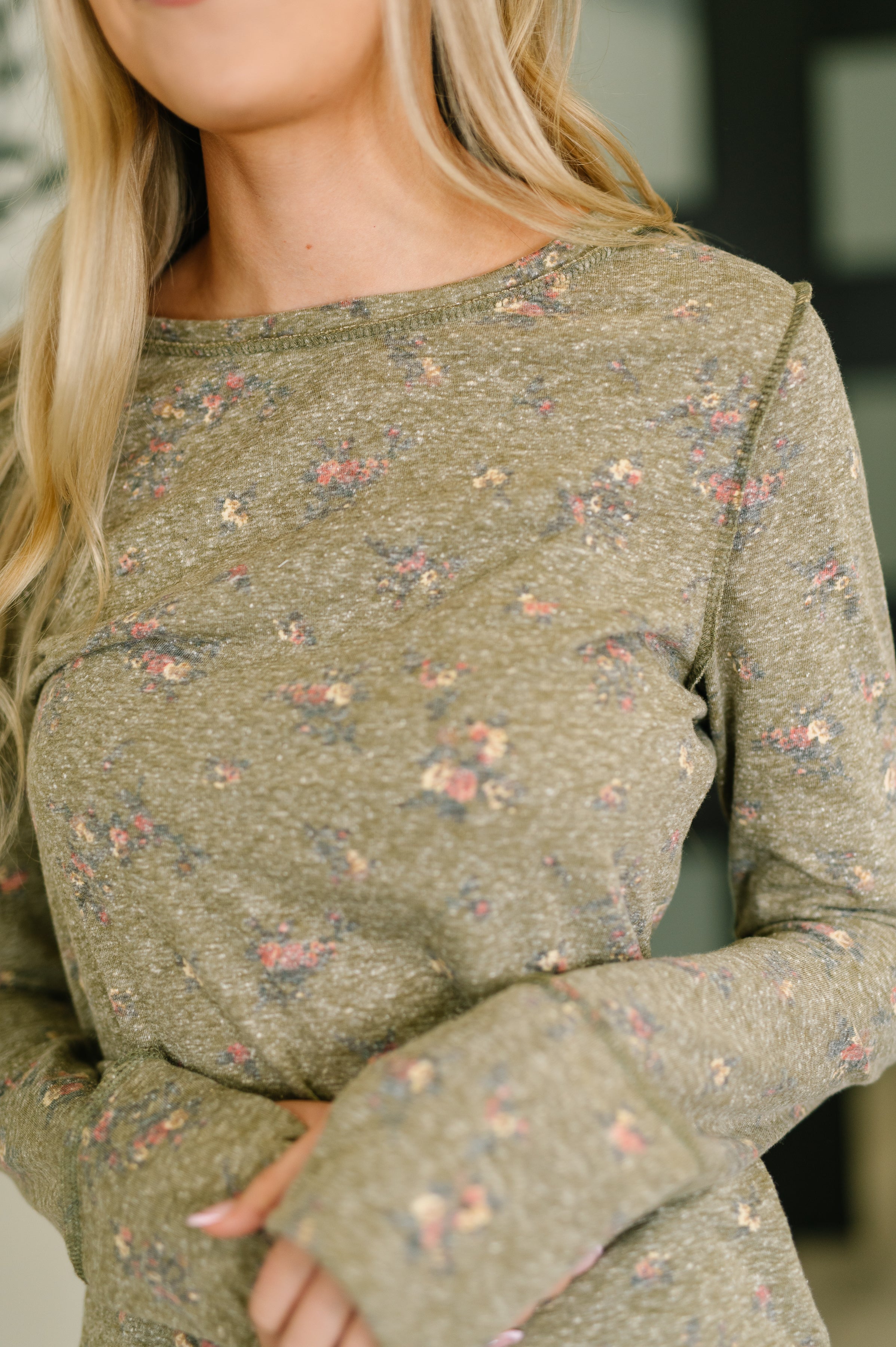 Close-up of woman wearing an olive long sleeve top with small scattered floral print and raw edge details indoors