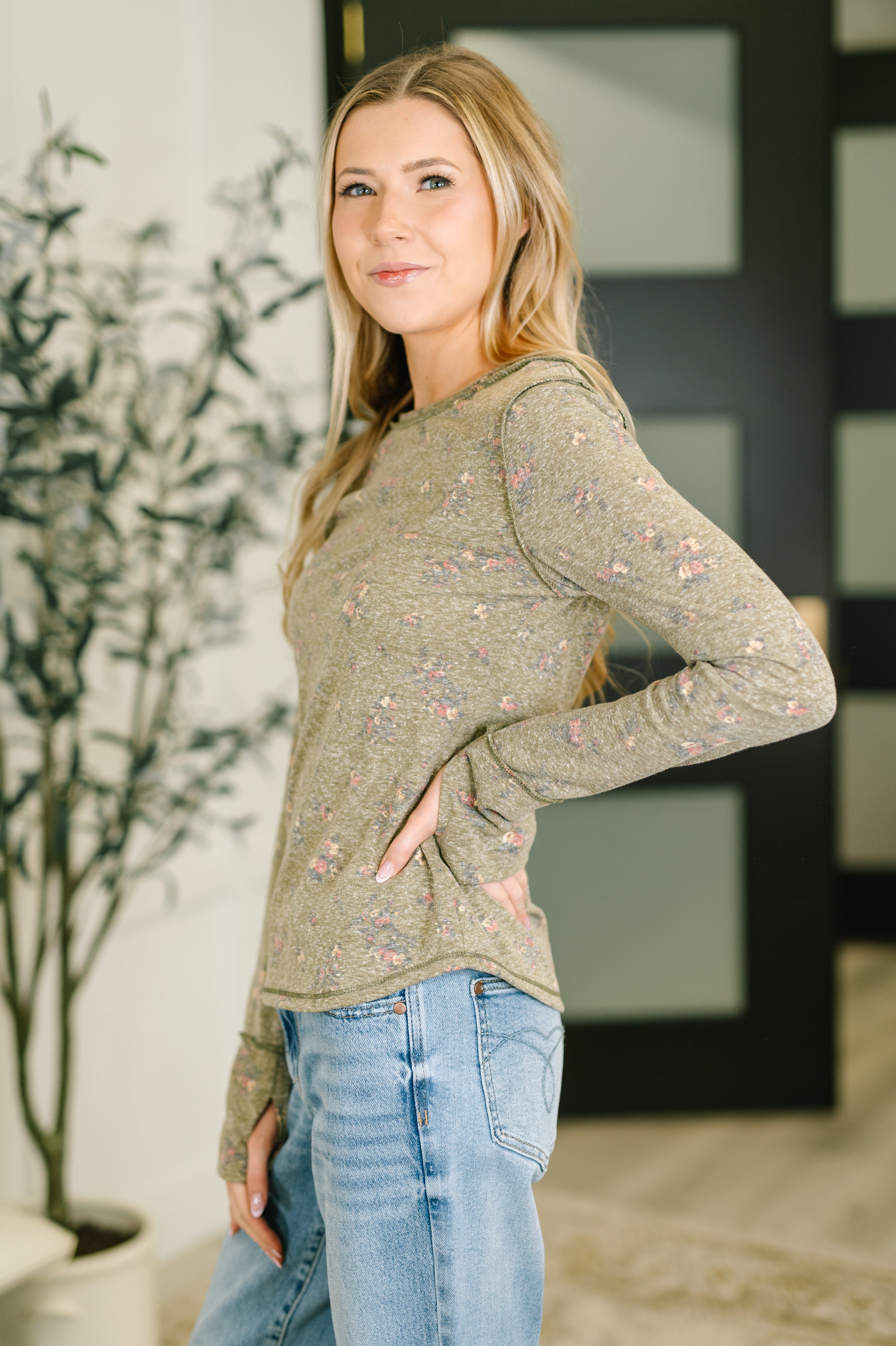 Woman wearing olive long sleeve top with subtle floral pattern paired with light blue jeans standing indoors