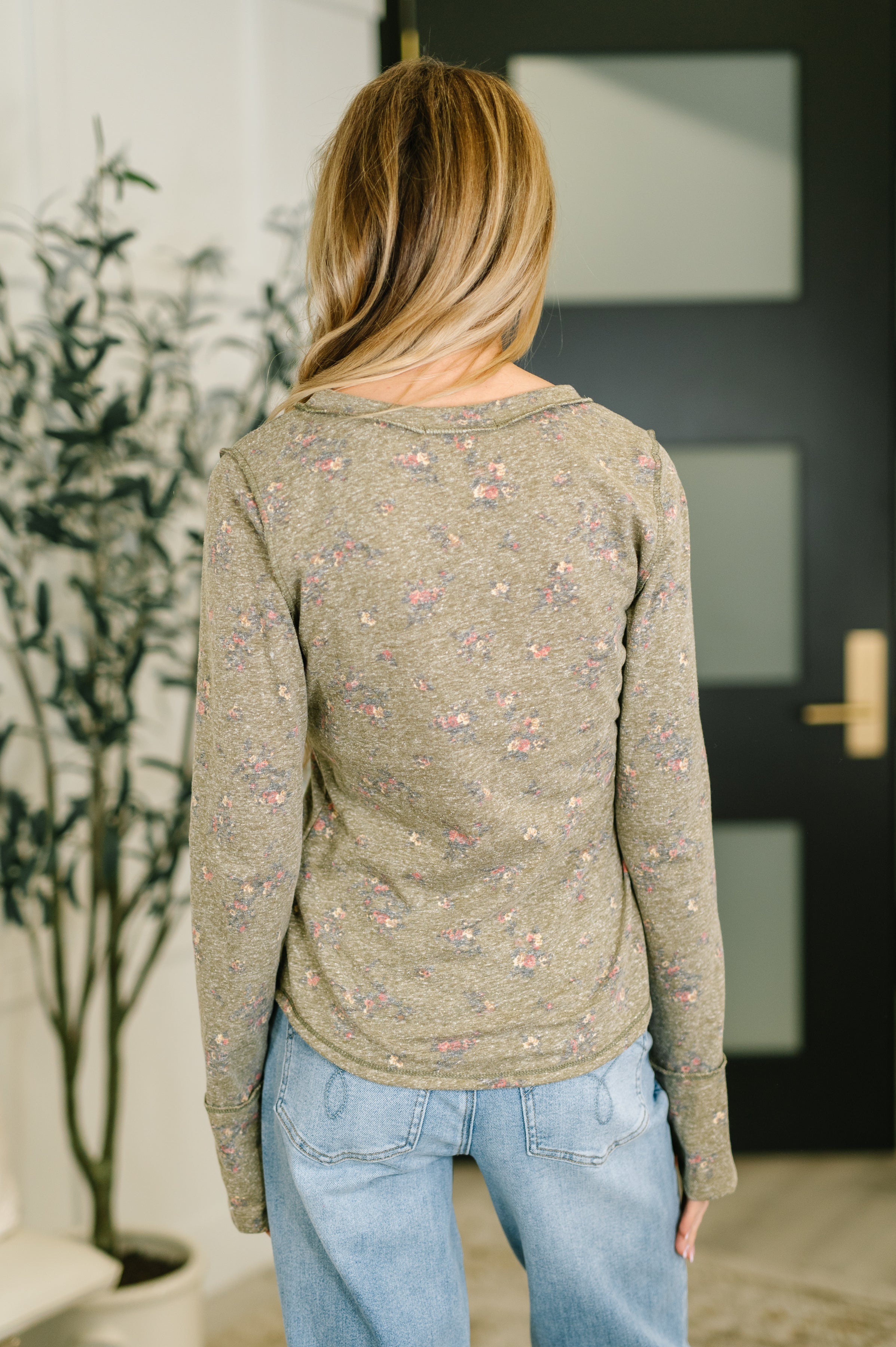 Woman wearing an olive long sleeve top with small floral print paired with light blue jeans standing indoors