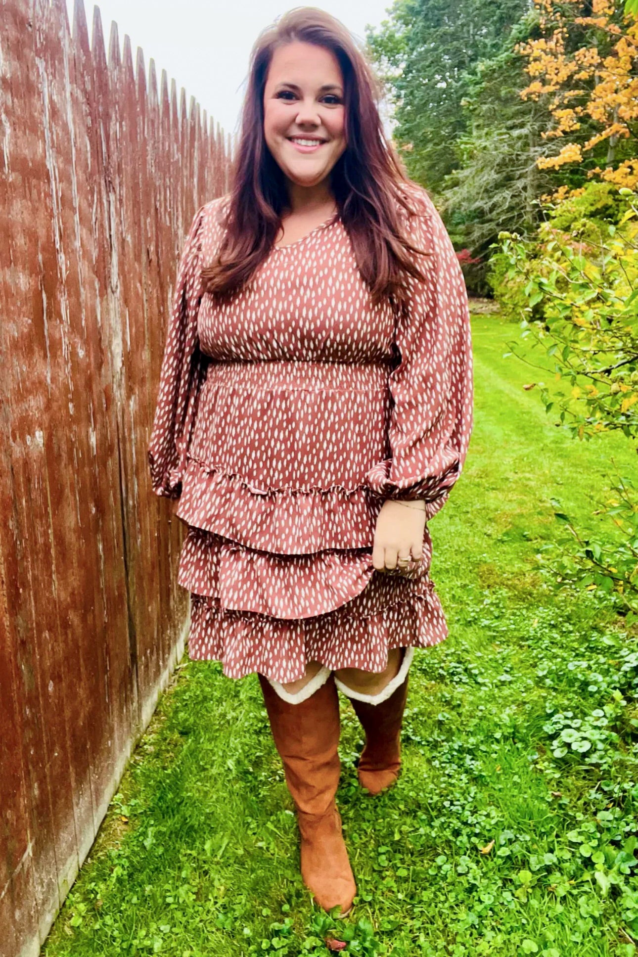 Woman smiling outdoors wearing a terracotta dress with white dash pattern, elastic waist, long sleeves, and layered ruffle skirt