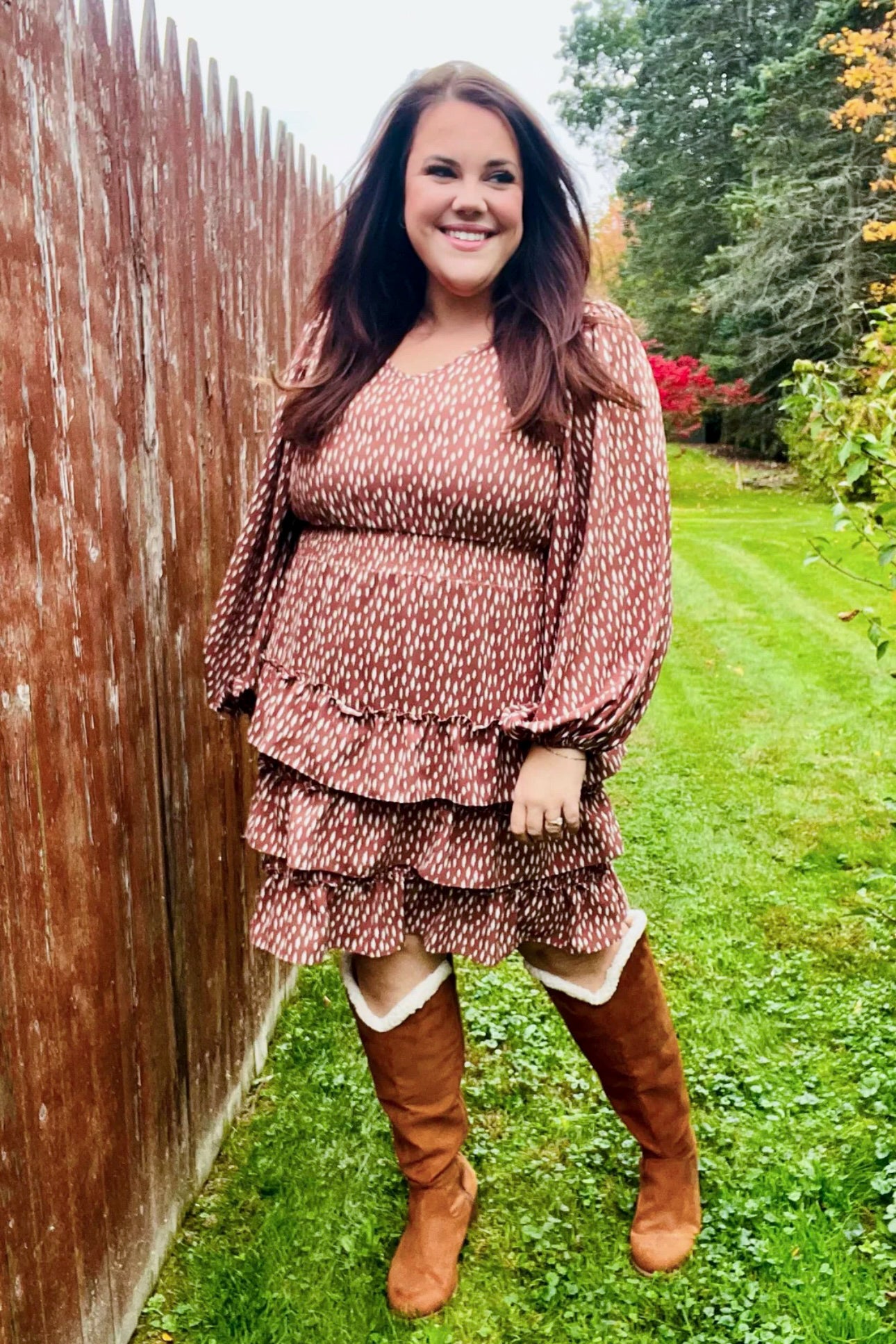 Smiling woman outdoors wearing a terracotta dress with white dash print, tiered ruffles, long voluminous sleeves, and knee-high boots
