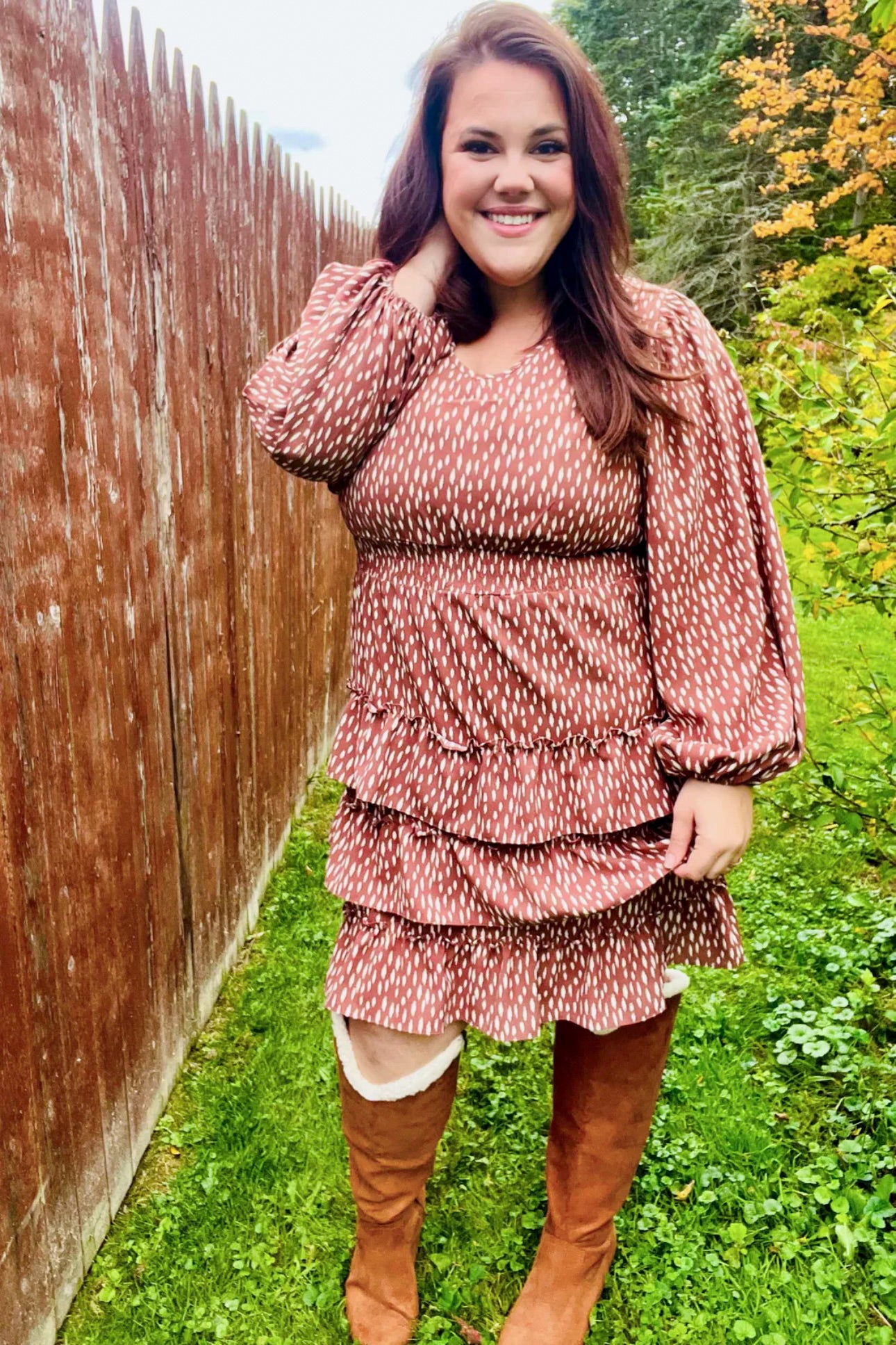 Woman smiling outdoors wearing a terracotta dress with white dash pattern, elastic waist, balloon sleeves, and layered ruffle skirt