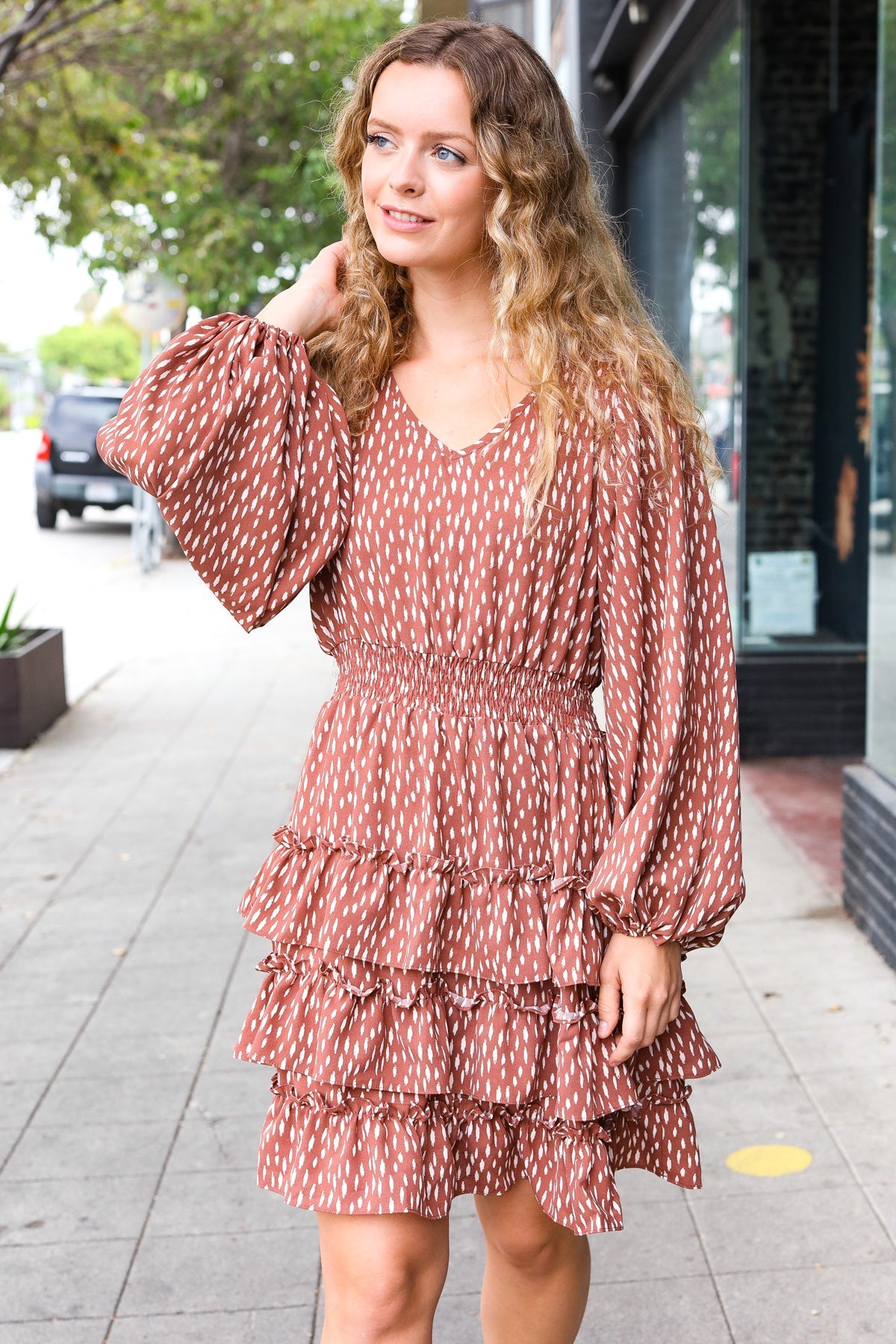 Woman wearing a terracotta dress with white abstract spots, tiered skirt, and voluminous long sleeves outdoors on sidewalk