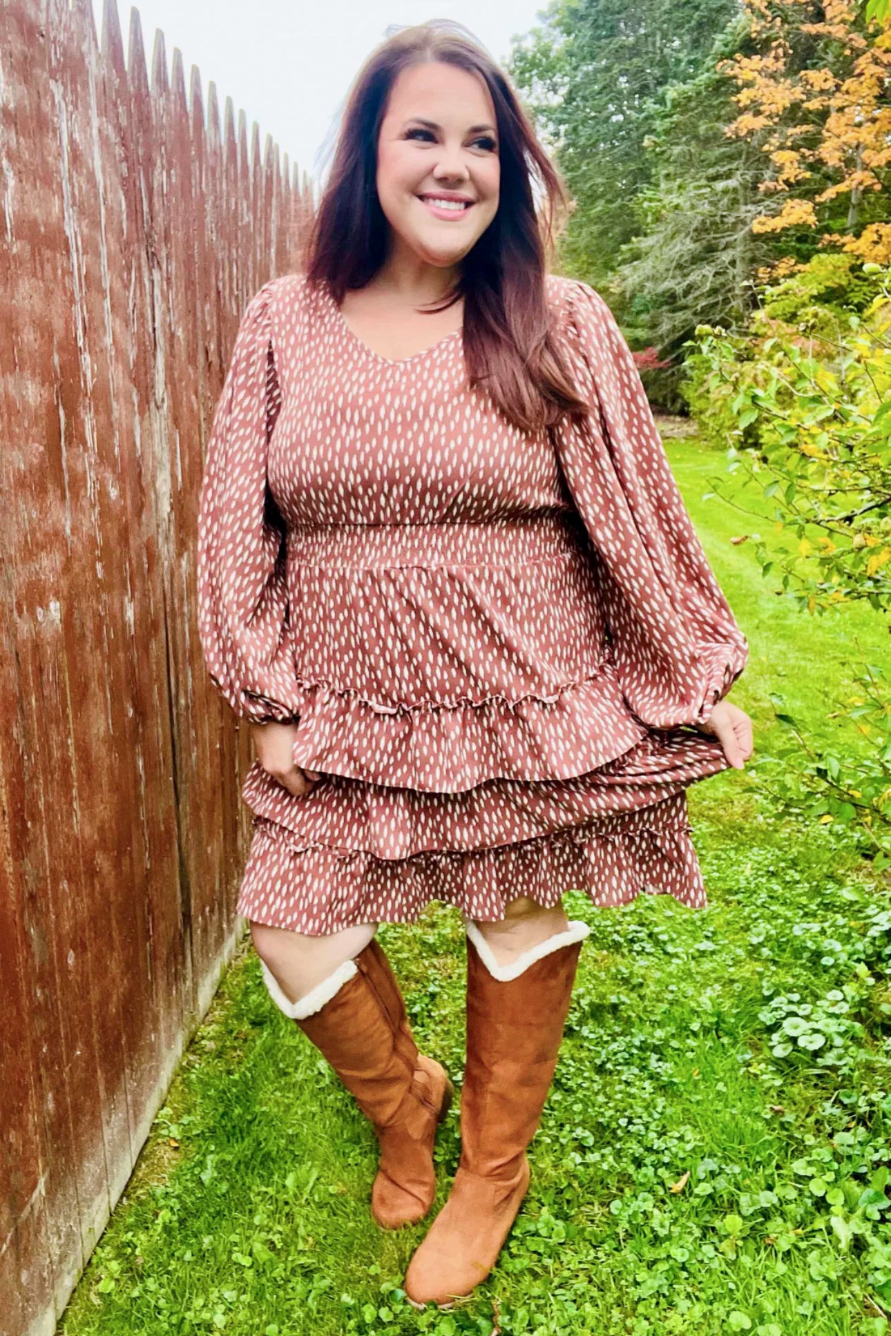 Smiling woman outdoors wearing a terracotta dress with white dash pattern, smocked waist, long puffed sleeves, and layered ruffle skirt