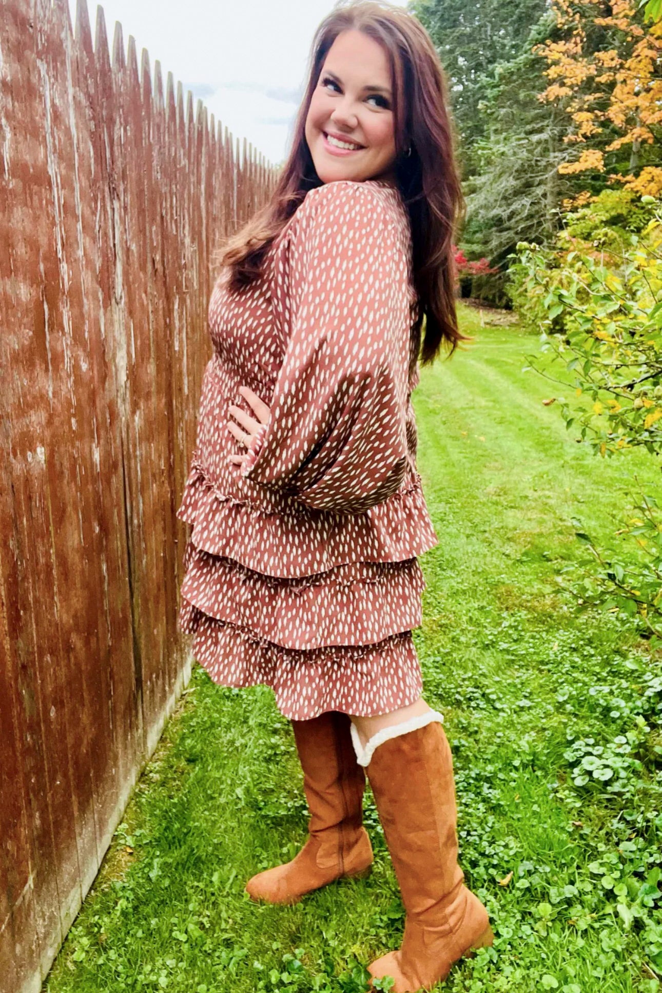 Smiling woman outdoors in a terracotta dress with white dash print, voluminous sleeves, tiered ruffle skirt, and knee-high boots