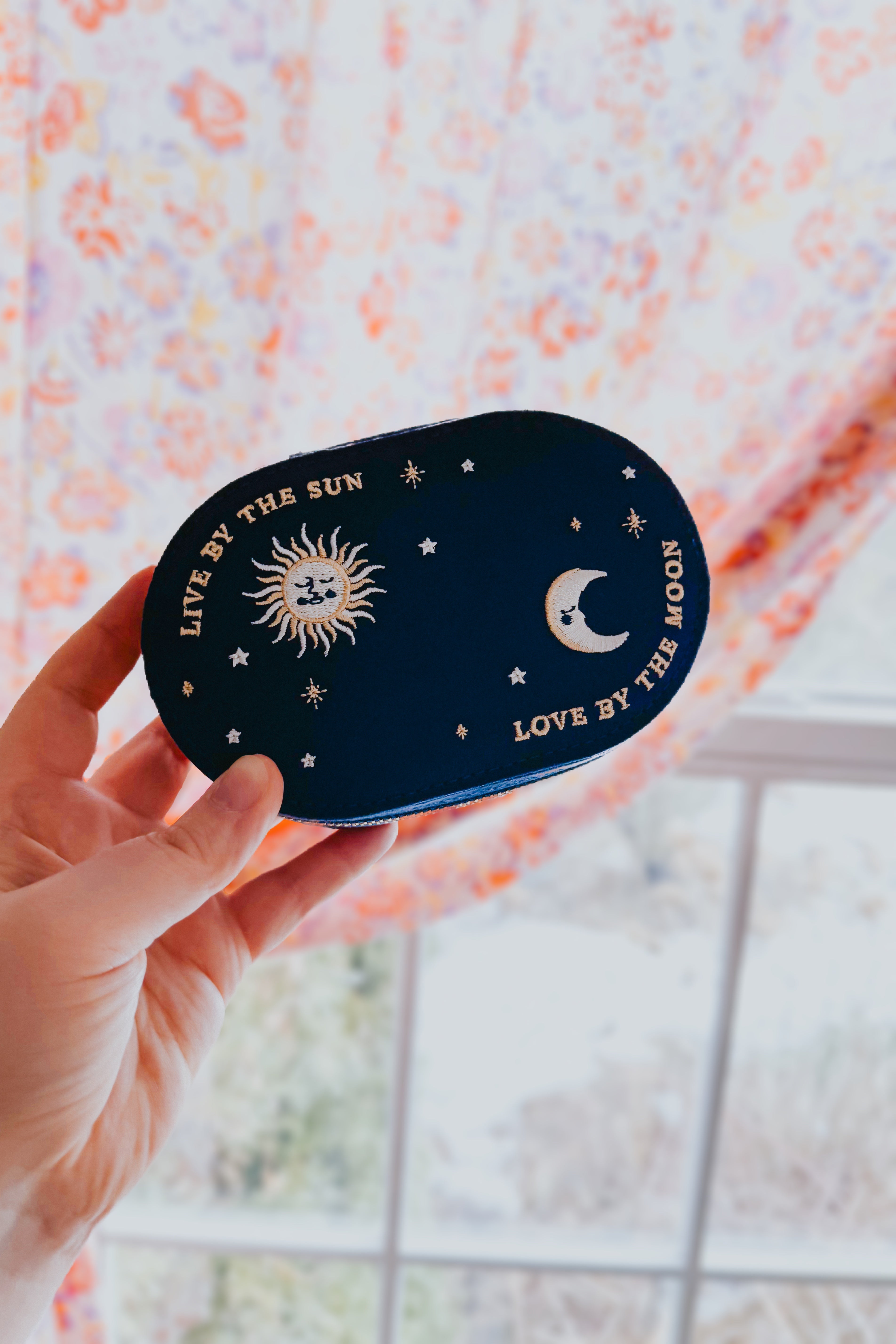 Navy-colored oval jewelry case with embroidered sun, moon, and stars and the phrases live by the sun love by the moon