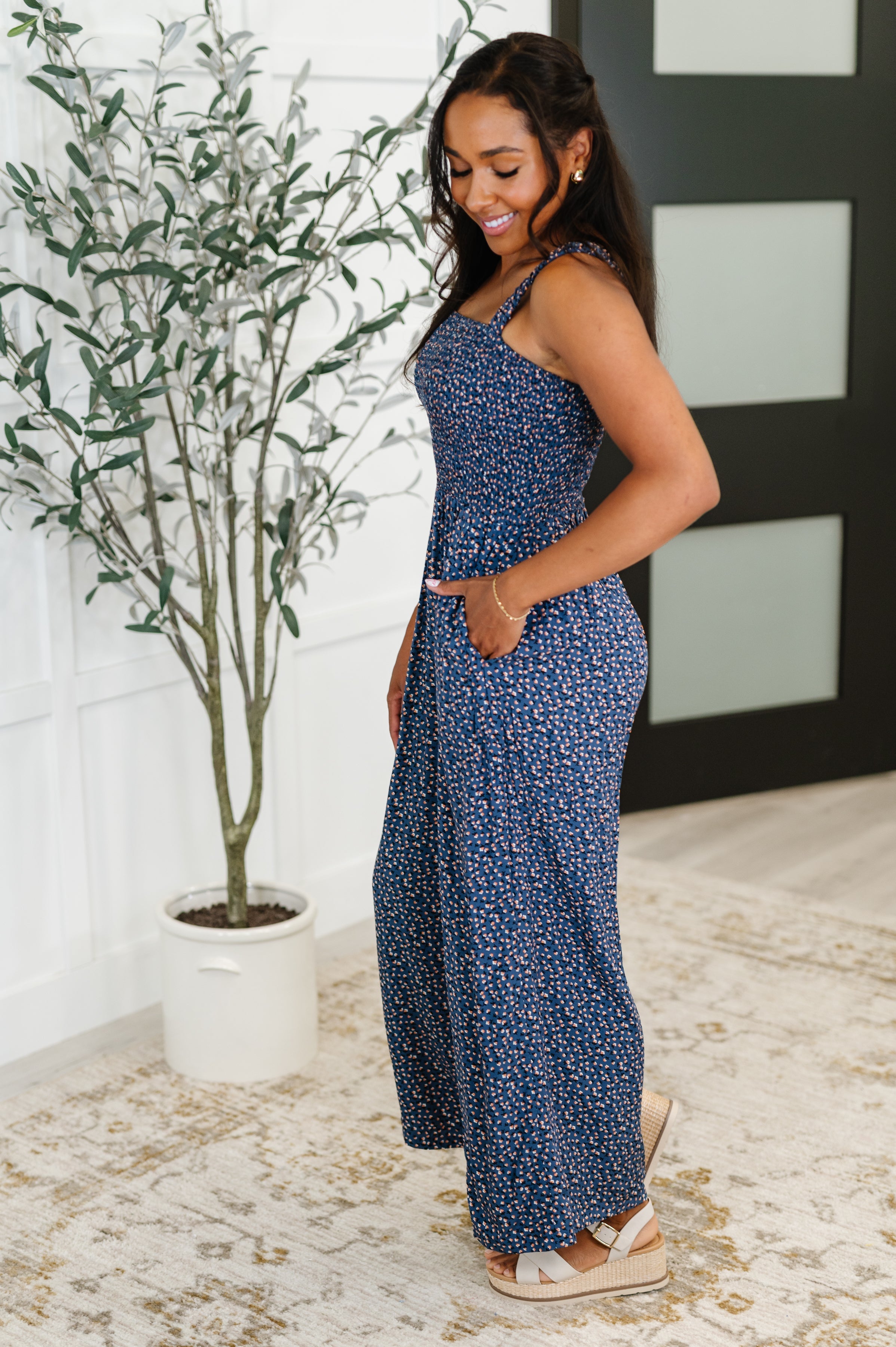 Woman smiling wearing a blue floral jumpsuit with smocked bodice and wide legs, standing indoors near a potted plant