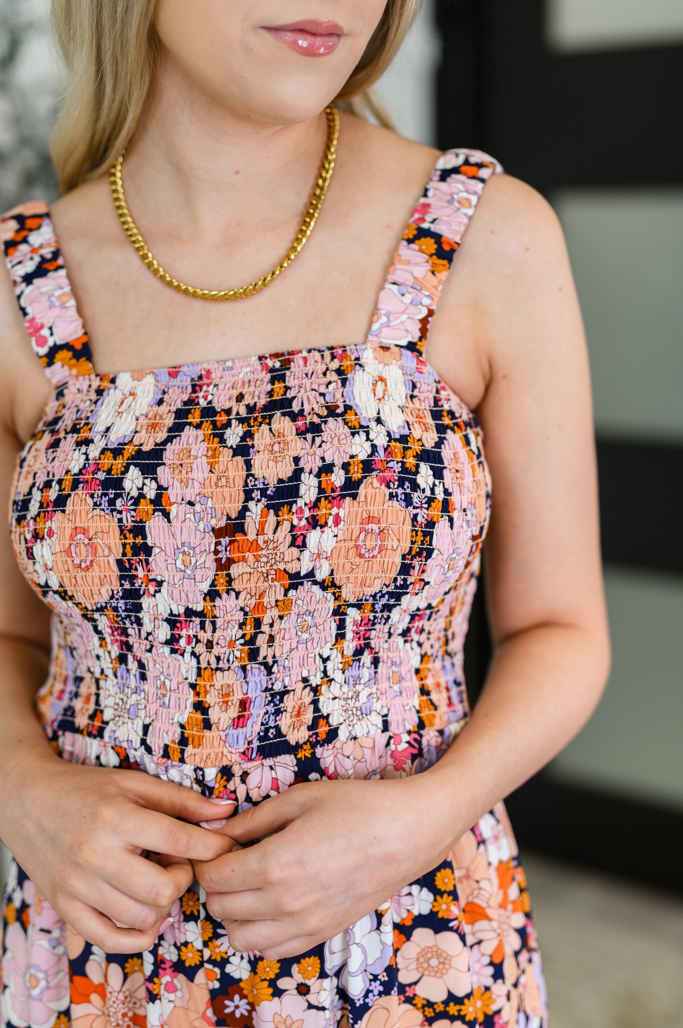 Close-up of a woman wearing a pink floral smocked top dress with wide straps and a gold chain necklace indoors