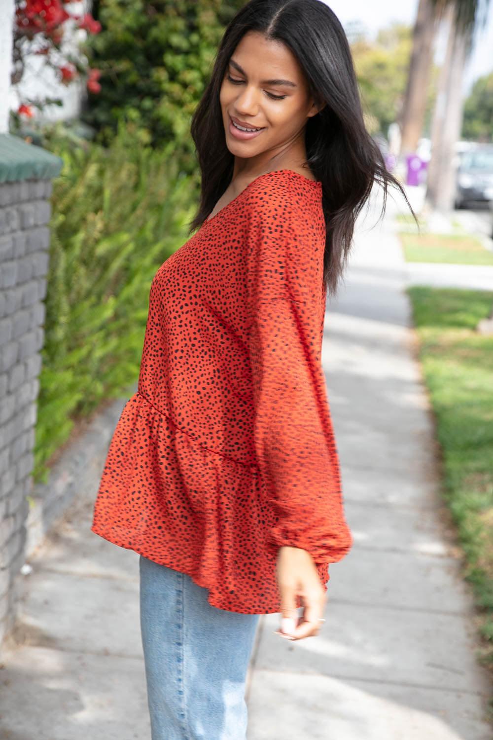 Woman outdoors wearing a rust-colored blouse with small black spots and flowy long sleeves paired with light blue jeans