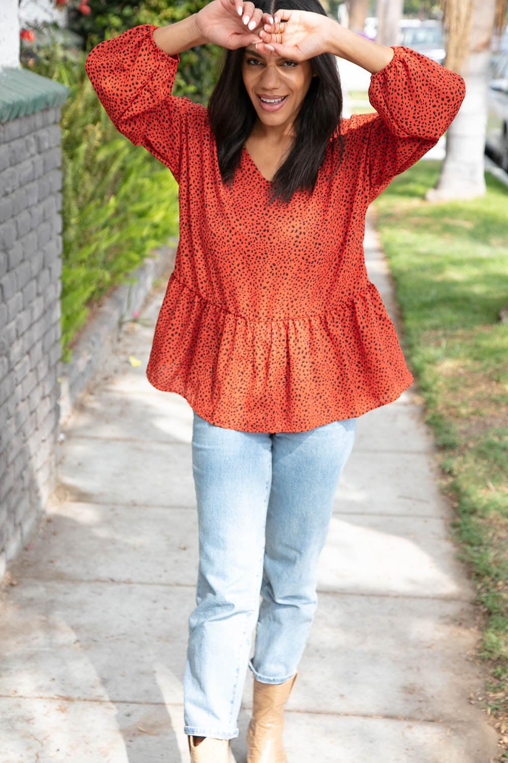 Woman outdoors wearing a rust blouse with small black spots and puffed three-quarter sleeves paired with light blue jeans
