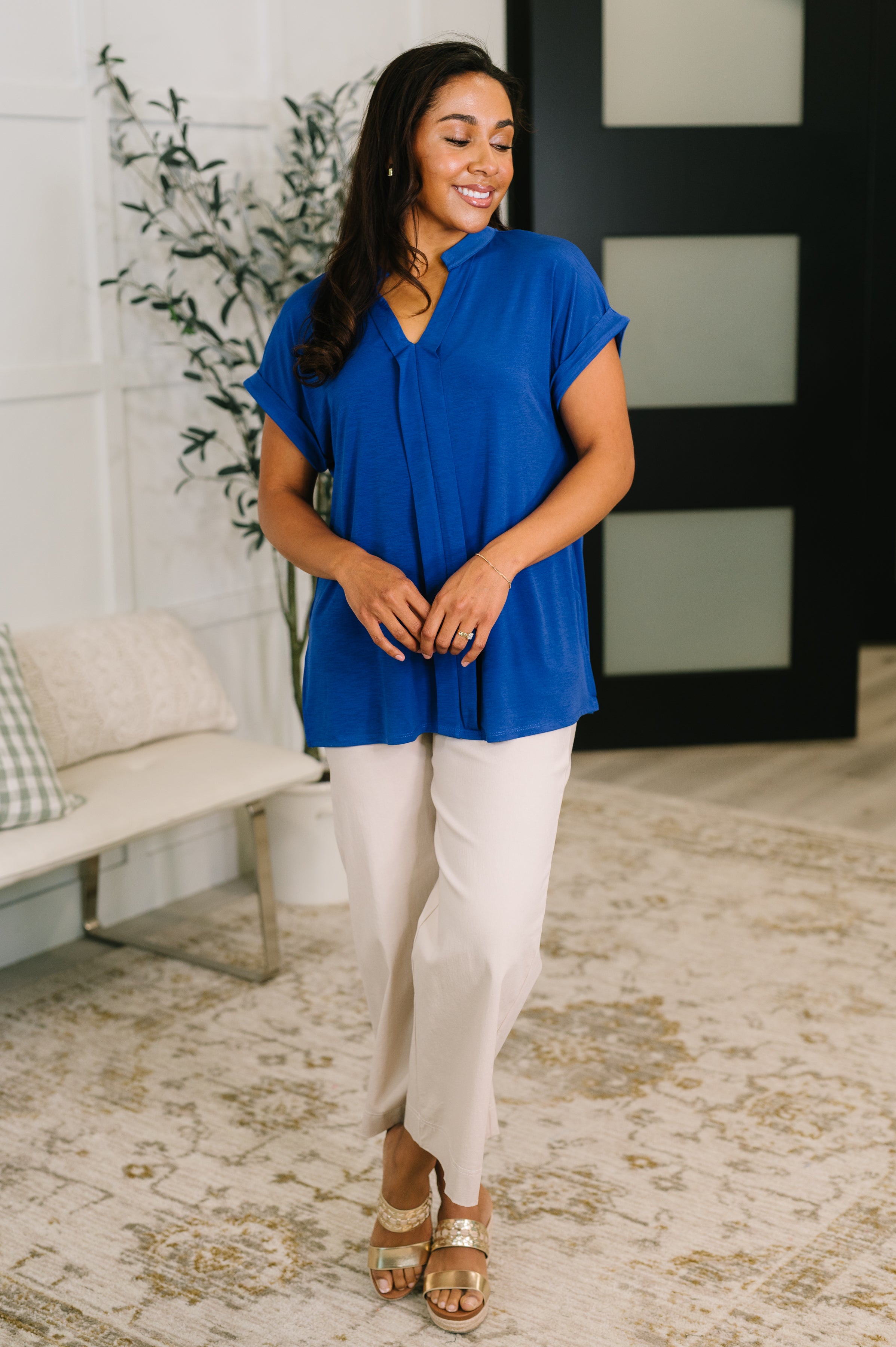 Woman wearing relaxed royal blue split neck blouse paired with light beige pants and sandals, smiling indoors