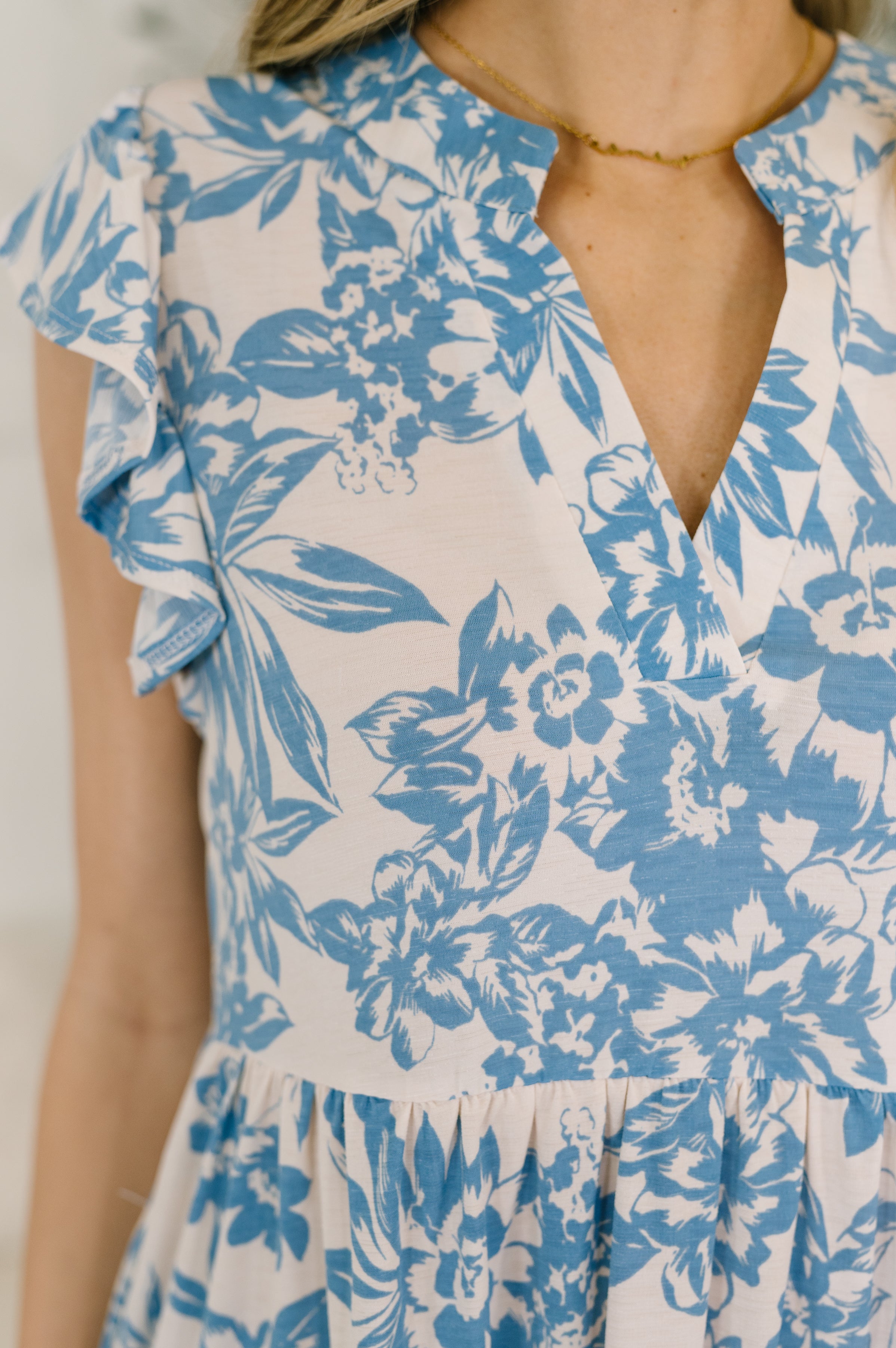 Close-up of a white dress with blue floral print, flutter sleeves, and a V-neckline on a woman indoors