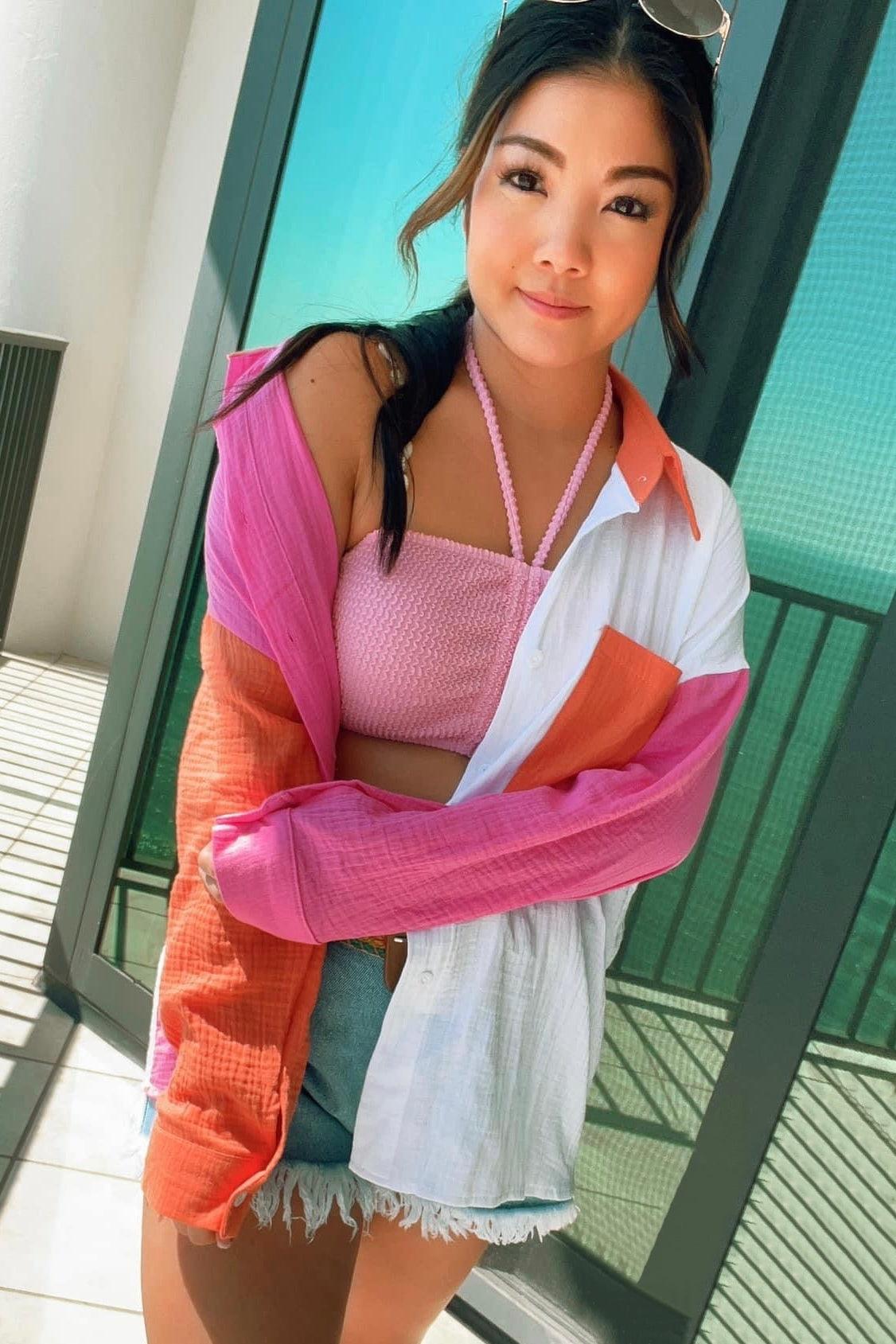 Woman wearing pink smocked crop top under pink, orange, and ivory color block shirt standing by balcony door in sunlight